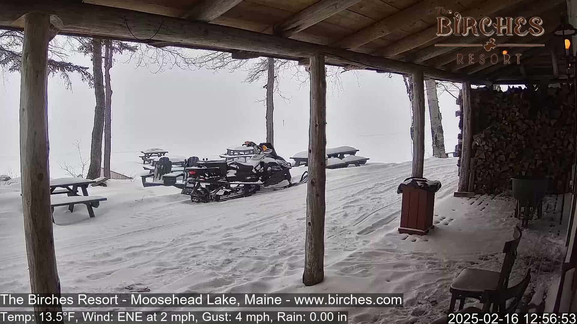 A snow-covered outdoor area with picnic tables, a snowmobile, and a woodpile is viewed from a covered porch in rainy, windy weather.