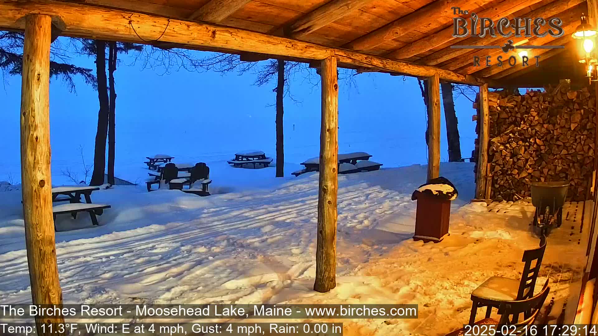 A snow-covered outdoor patio with picnic tables is viewed from inside a wooden structure under a twilight sky.