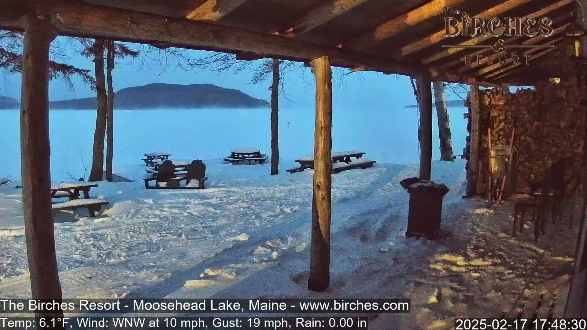A snow-covered picnic area with several empty tables is seen from under a wooden shelter overlooking a calm, misty lake and distant mountains in the cool twilight.