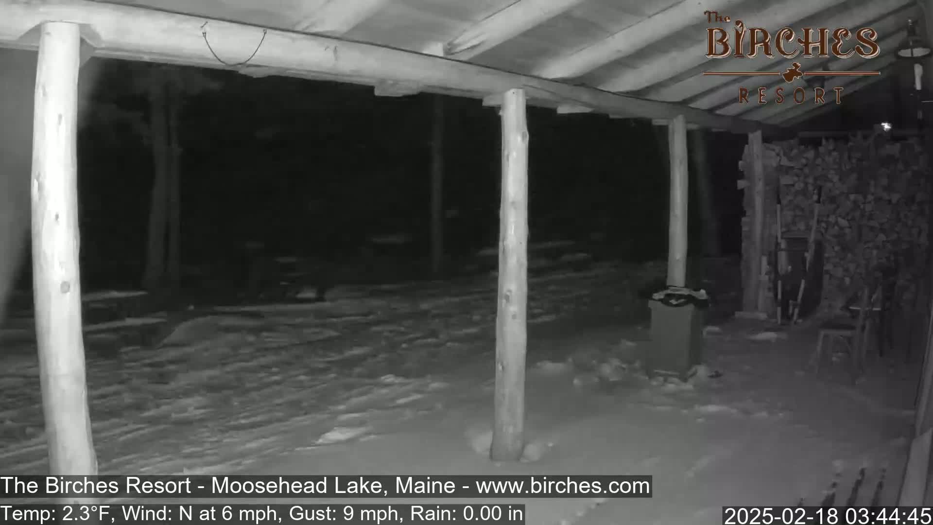A snow-covered porch area at night, under a wooden structure, with a woodpile visible in the background, a north wind of 6 mph gusting to 9 mph, and a temperature of 2.3°F.