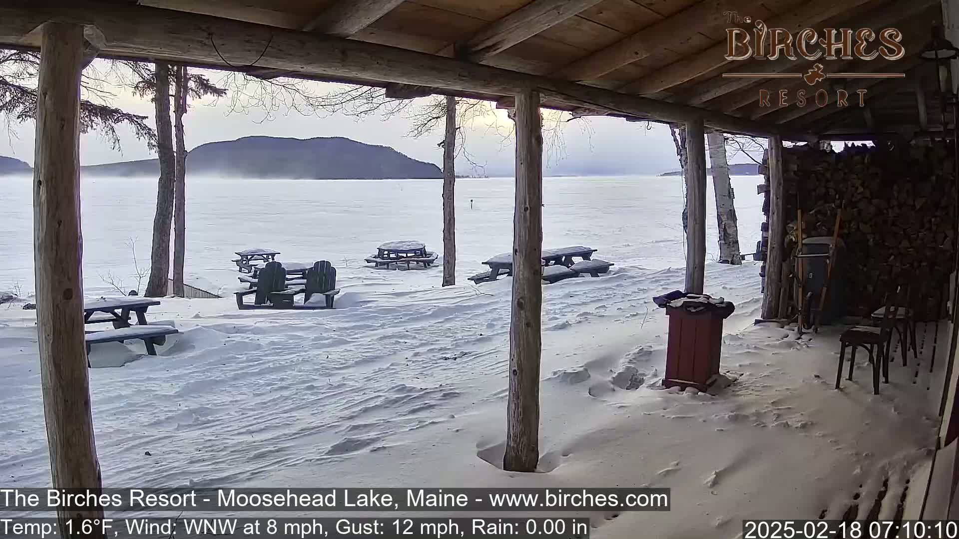 A snow-covered lakeside scene viewed from a wooden porch, showing picnic tables and a distant, hazy mountain range under a cloudy sky.
