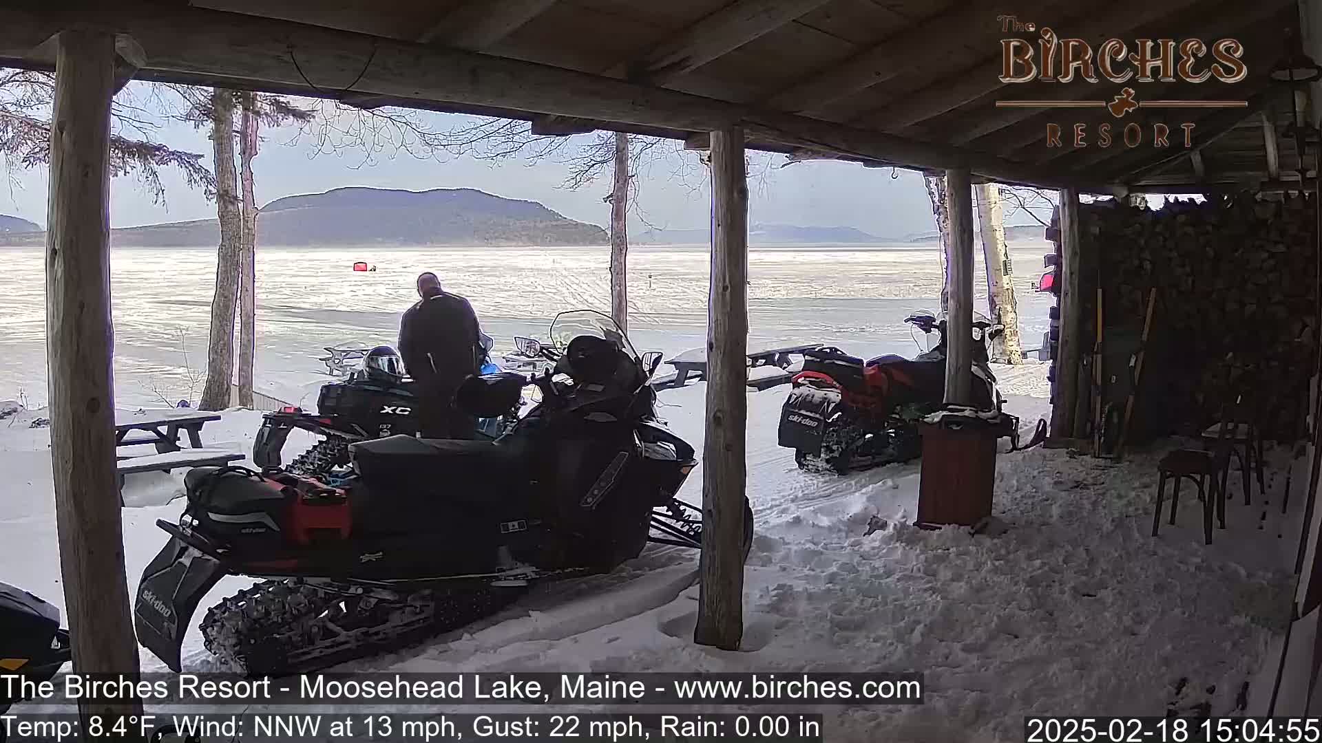 Under a covered porch, snowmobiles are parked in the snow overlooking a frozen lake and distant mountains on a windy, cold day.