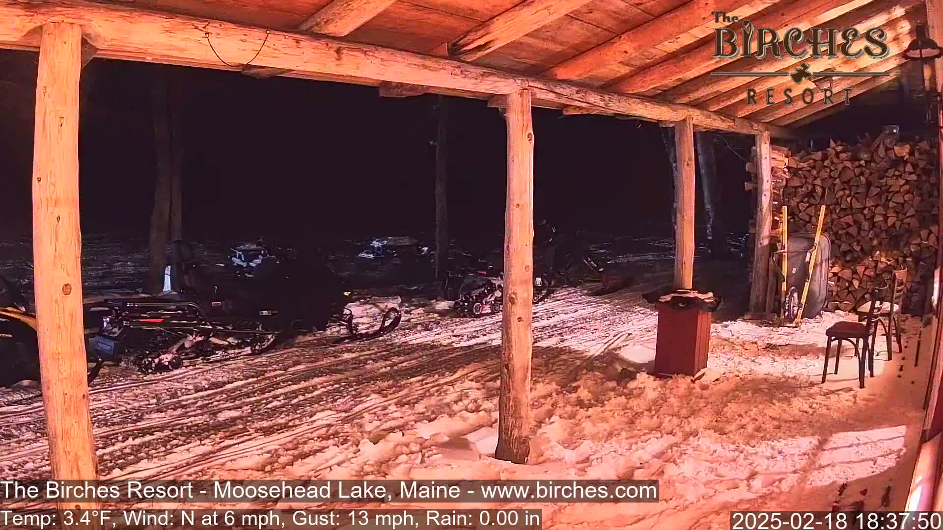 Snowmobiles are parked outside a rustic wooden structure at night in snowy conditions with a north wind of 6 mph and gusts up to 13 mph.