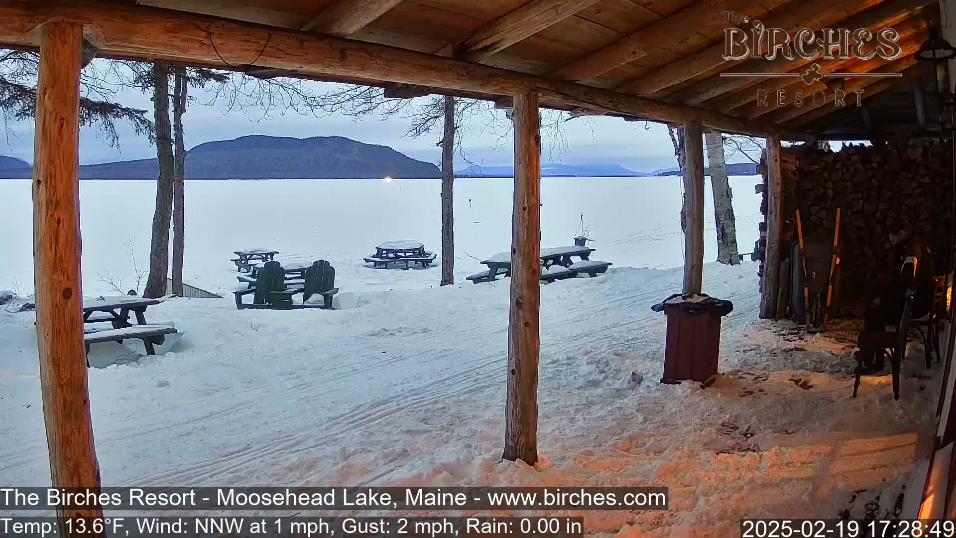A snow-covered lakeside view is seen from a wooden porch with picnic tables and chairs, under a cloudy sky.