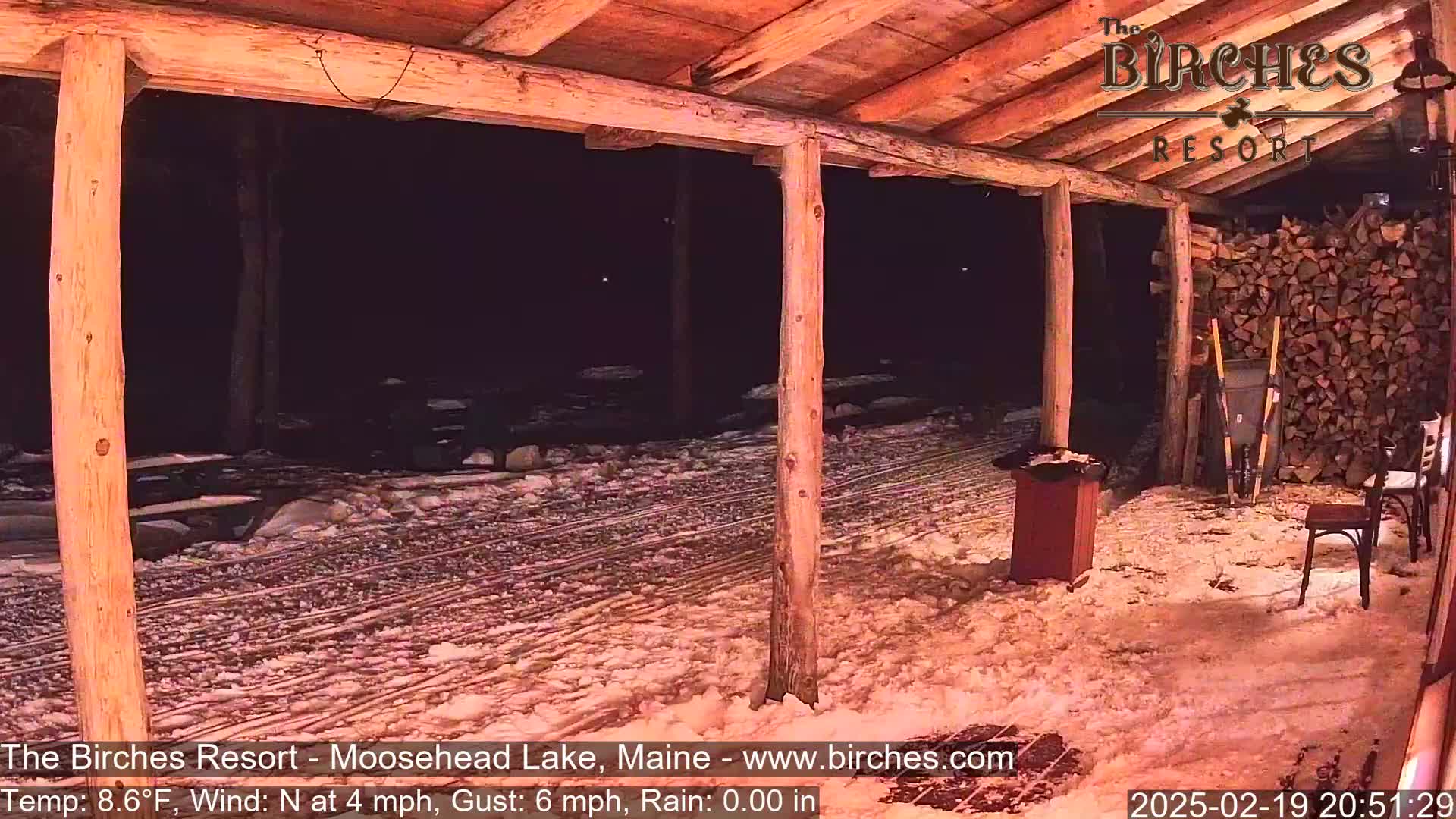 A snow-covered outdoor patio area at night, viewed from under a wooden structure, with a woodpile visible in the background.