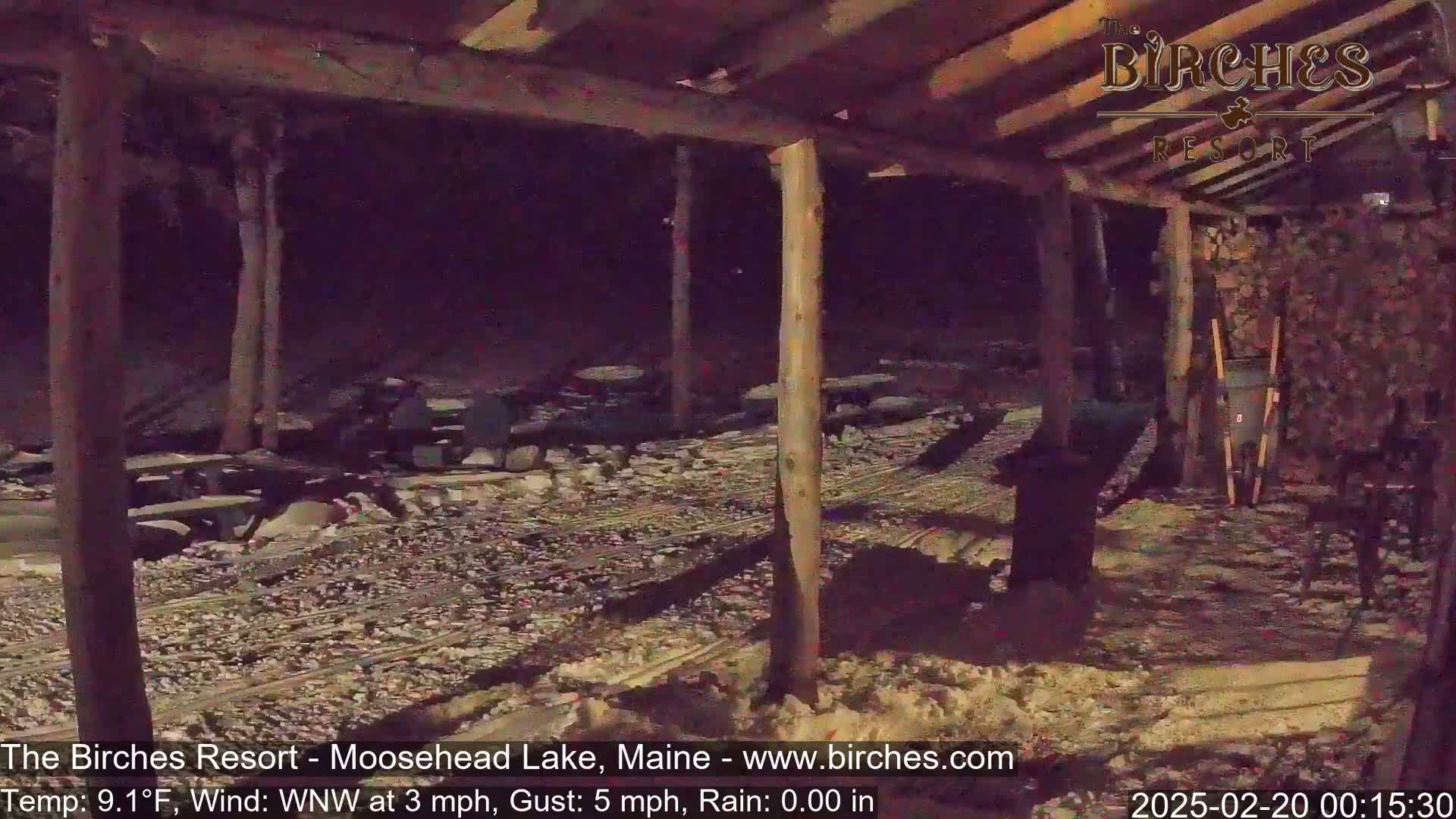 A snow-covered outdoor area at night, partially sheltered by a wooden structure with picnic tables, is seen under a 9.1°F temperature with a WNW wind at 3 mph and gusts up to 5 mph.