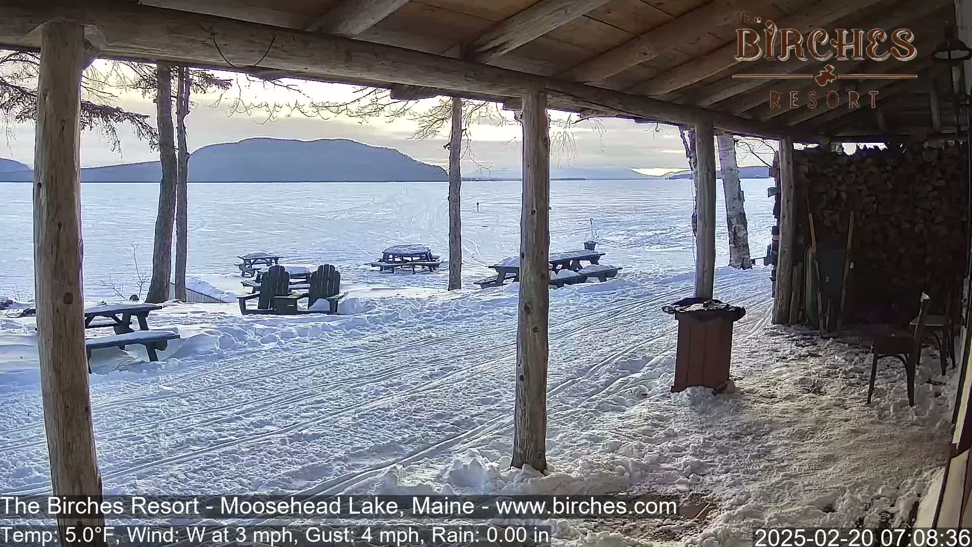 A snow-covered lakeside patio with picnic tables and chairs is viewed from a wooden porch, overlooking a frozen lake and distant mountains under a partly cloudy sky.