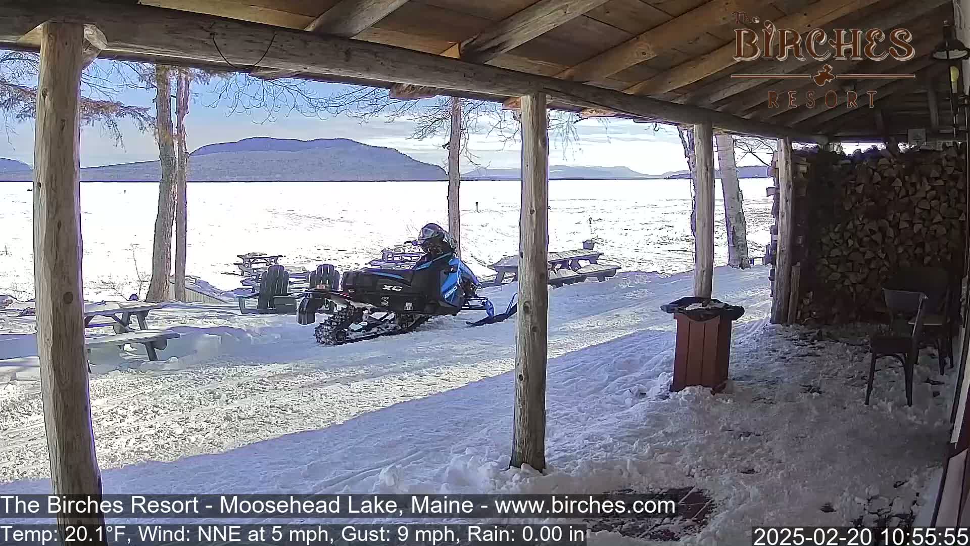 A snowmobile is parked on a snow-covered porch overlooking a frozen lake and distant mountains under a sunny sky.