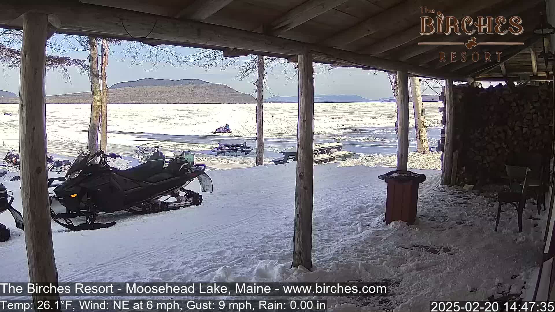 Snow-covered ground with snowmobiles parked near picnic tables under a wooden structure overlooks a partially frozen lake on a sunny, windy day with light rain.