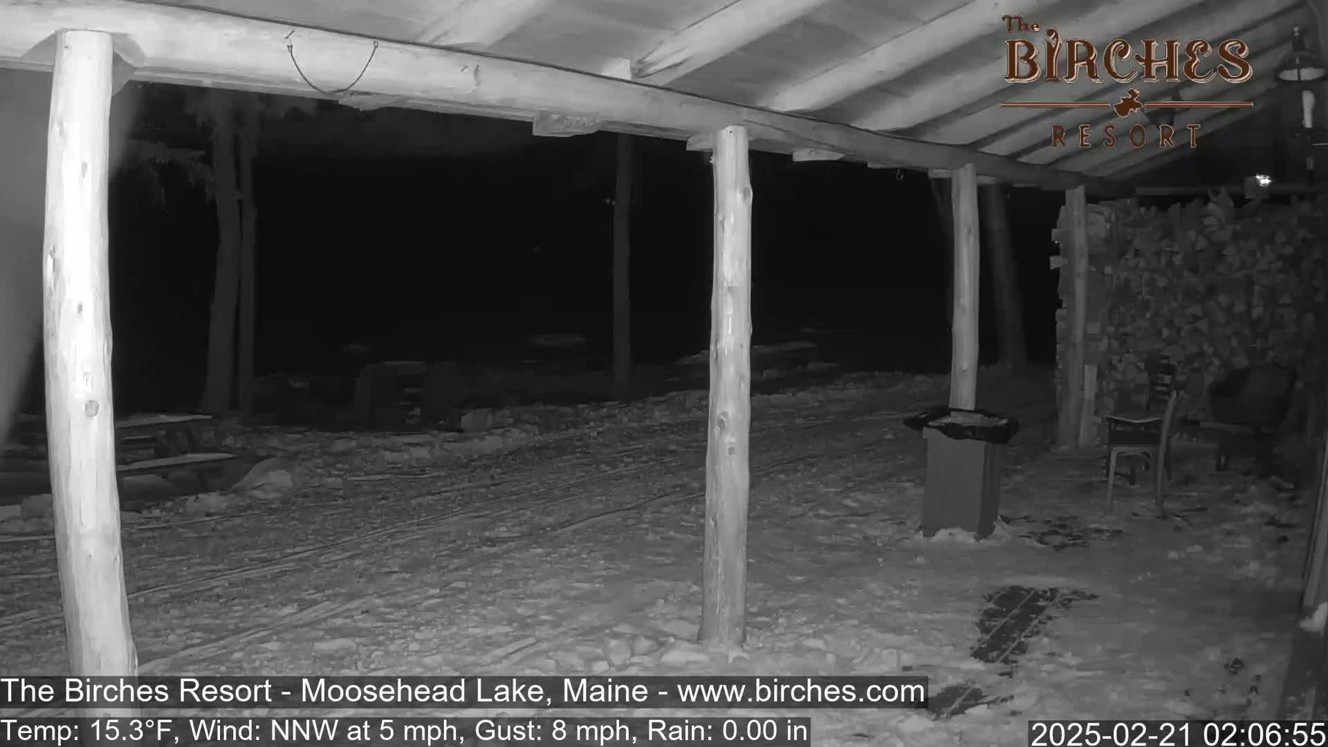 A snow-covered outdoor area at night, under a wooden covered porch, with a woodpile and chairs visible in the background, under conditions of 15.3°F, a north-northwest wind at 5 mph with gusts up to 8 mph, and no rain.
