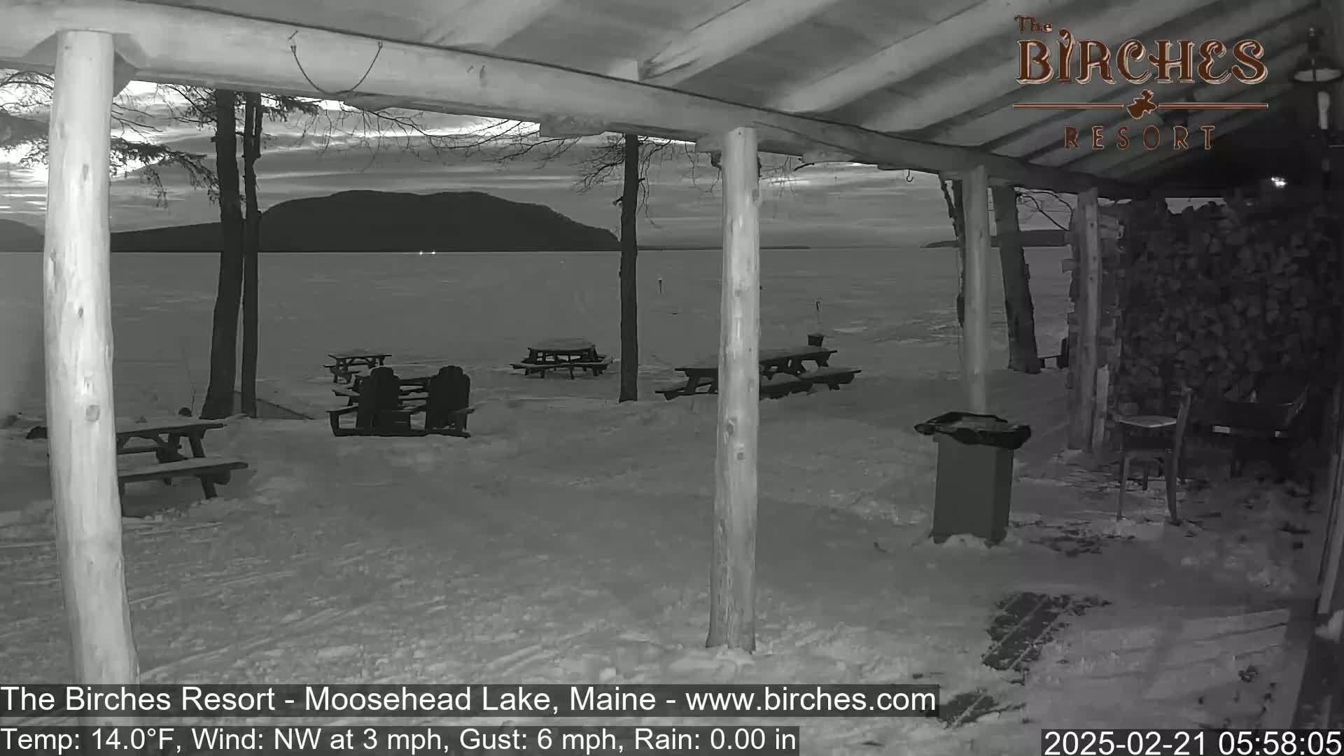 A snow-covered lakeside picnic area is viewed from a covered porch at dawn, with a calm lake and distant mountains visible in the background.
