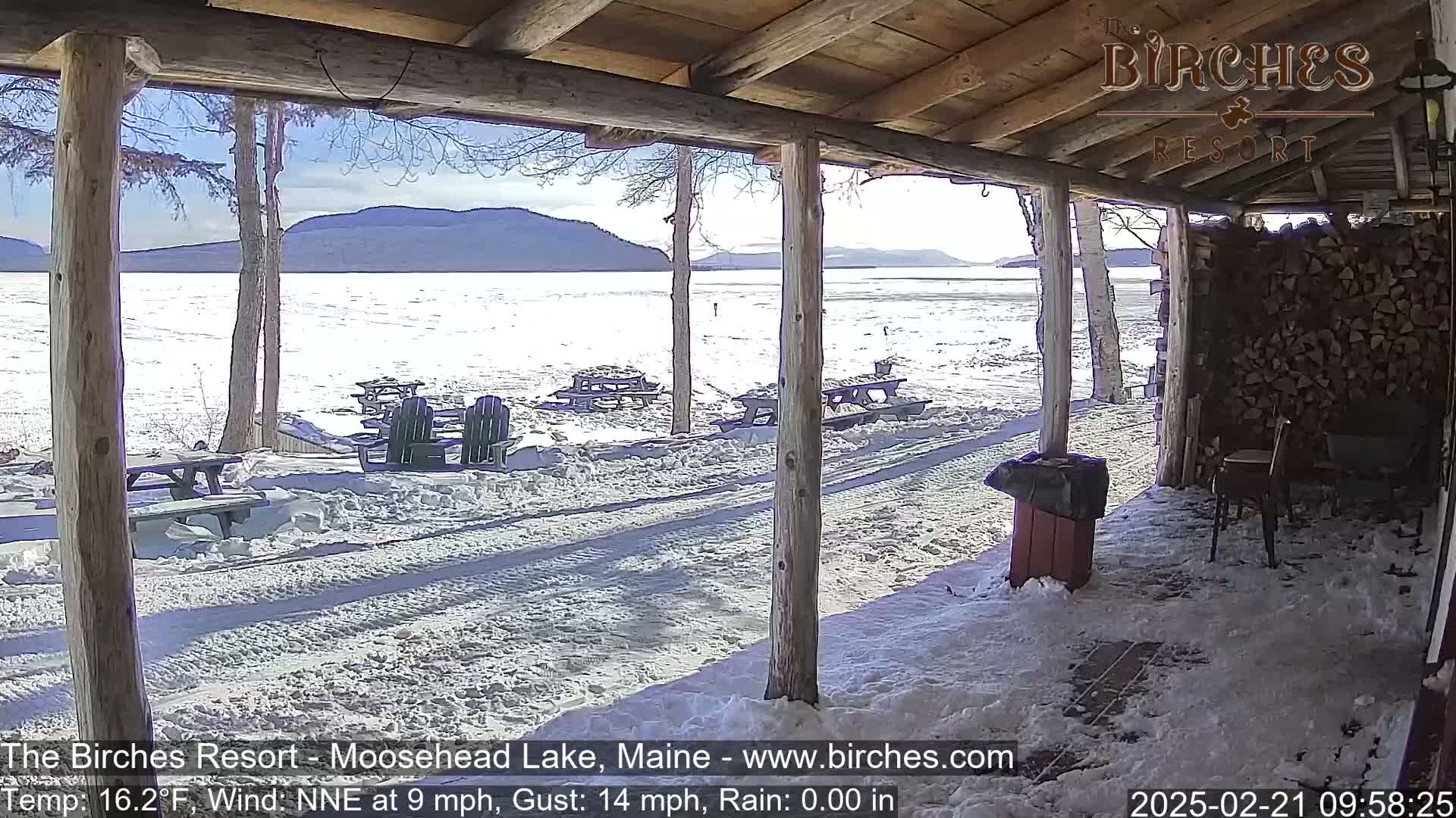 A snow-covered porch overlooks a frozen lake and distant mountains under a sunny sky.