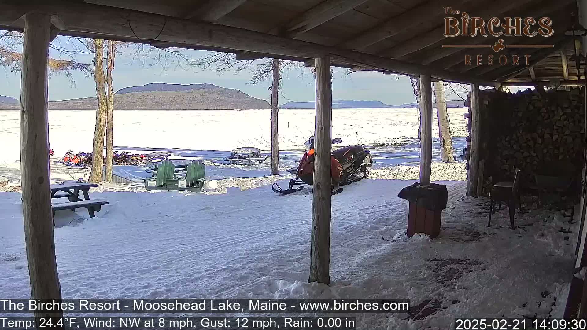 From a snow-covered porch, a view of a frozen lake with snowmobiles parked by the shore is visible under a sunny sky.