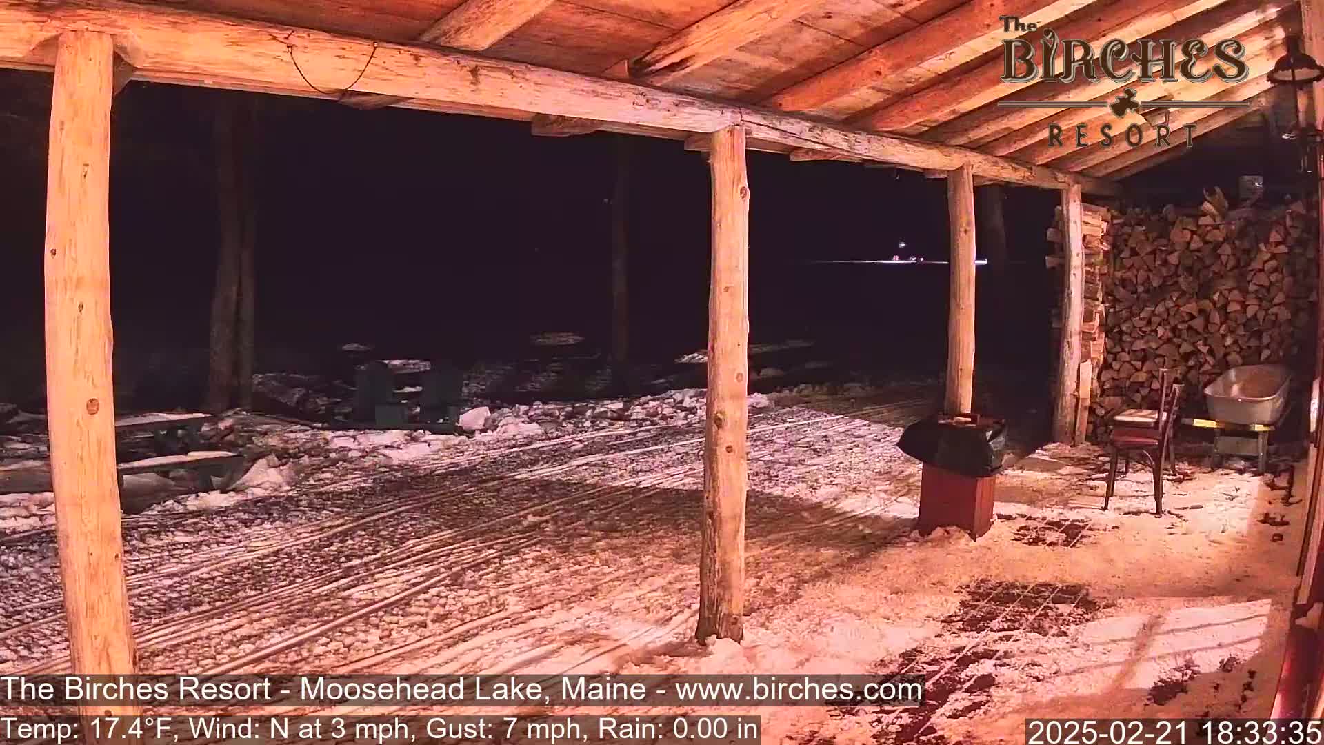A snow-covered outdoor patio area with wooden posts and picnic tables is visible at night under a partly cloudy sky, with a gentle north wind.