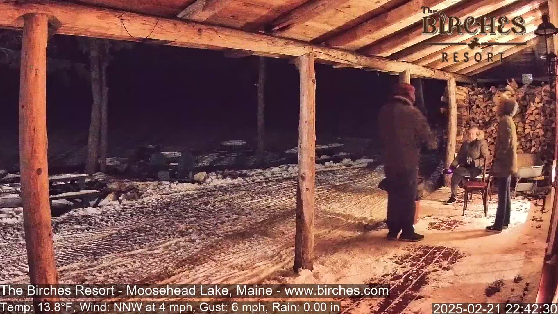 Under a wooden shelter at night in snowy conditions, three people stand on a snow-covered ground near a woodpile.