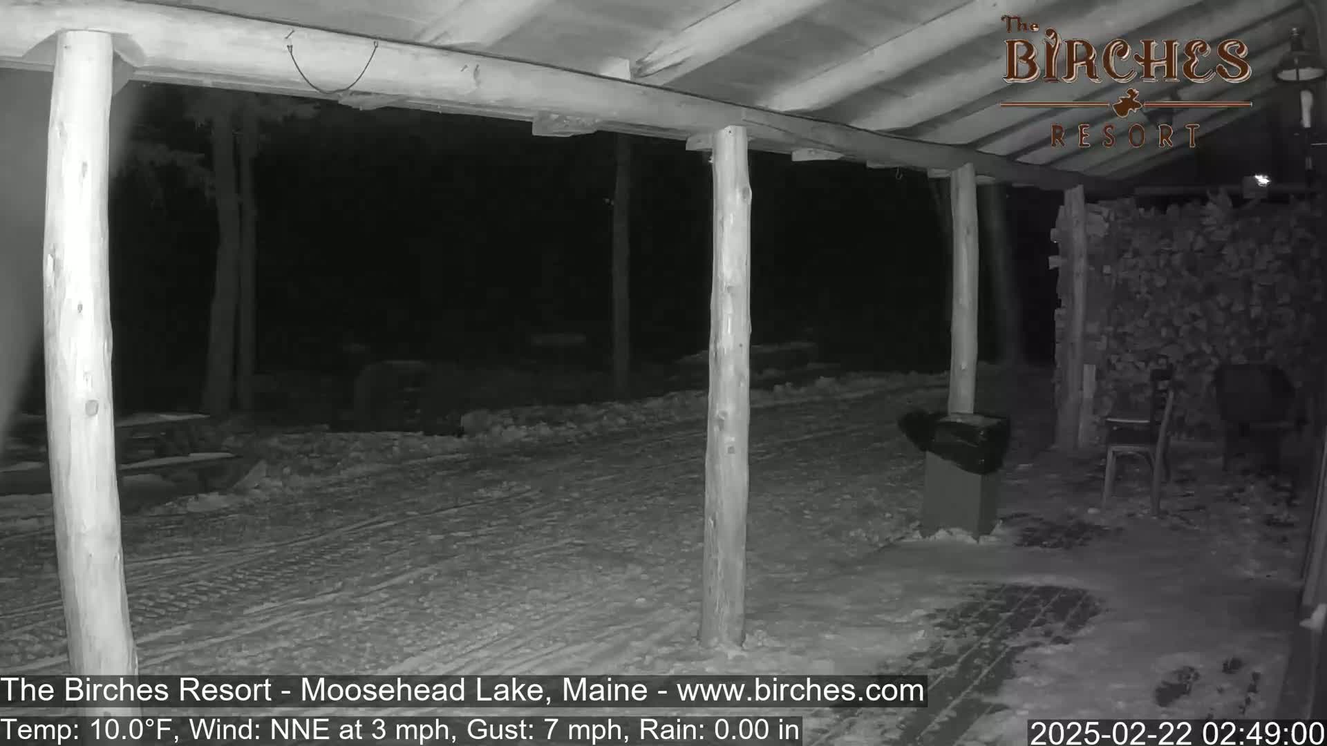 A snow-covered outdoor area at night, viewed from under a wooden porch, with a wind from the north-northeast at 3 mph and gusts up to 7 mph.