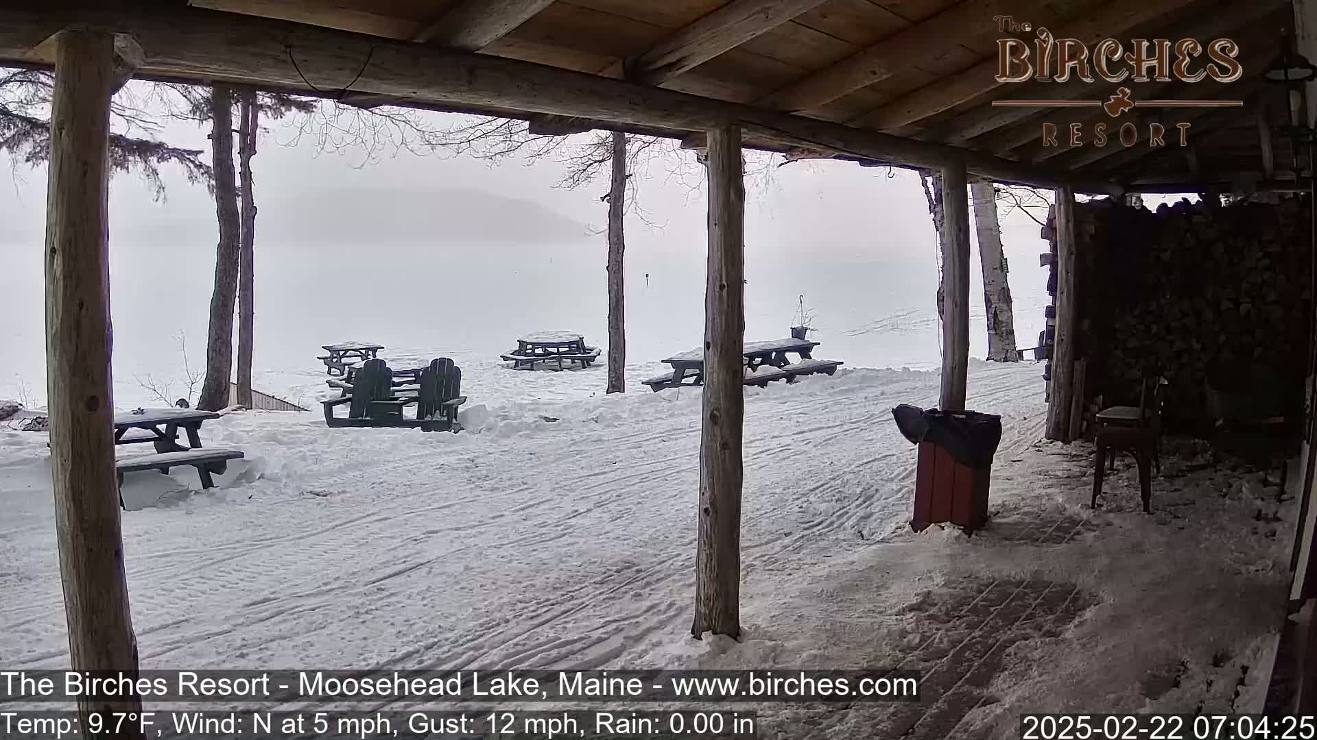 A snow-covered outdoor area with picnic tables and Adirondack chairs is seen from a covered porch on an overcast day.