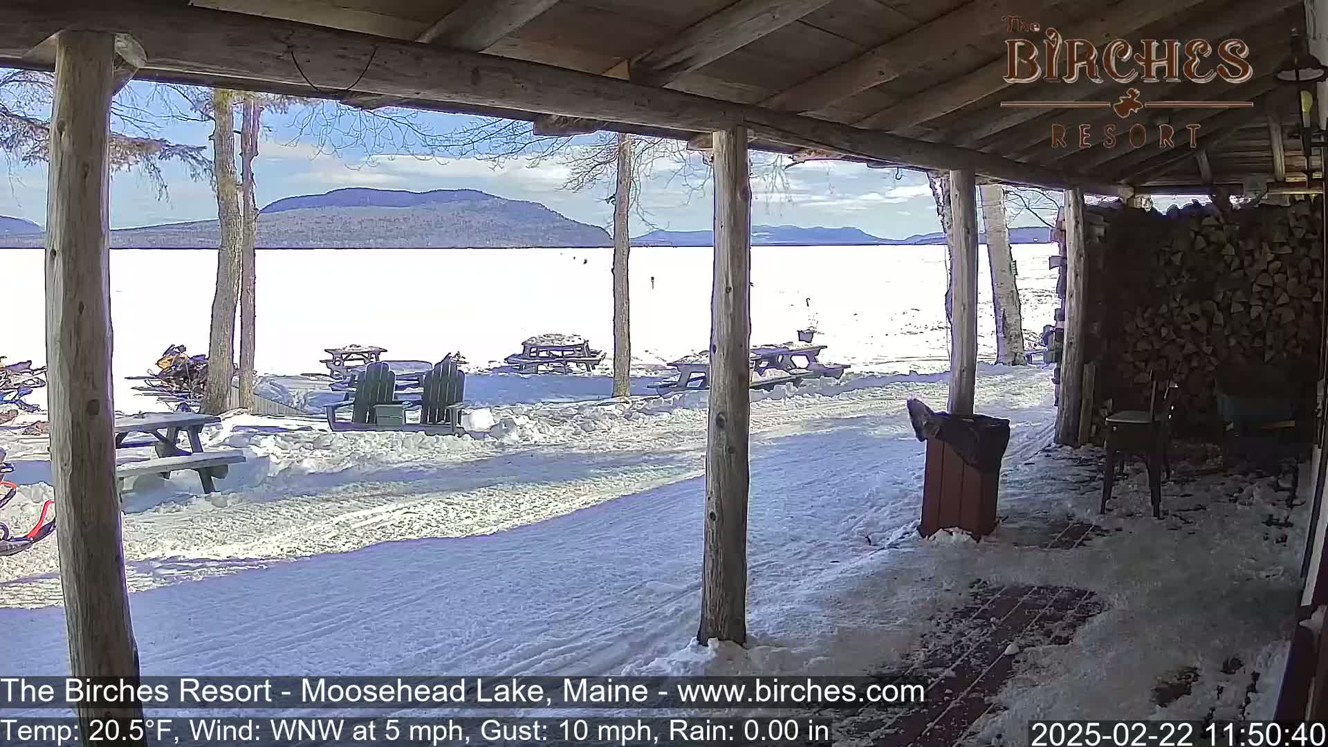 A snow-covered lakeside patio with picnic tables and snowmobiles is seen from a covered porch on a partly sunny day with a light wind.