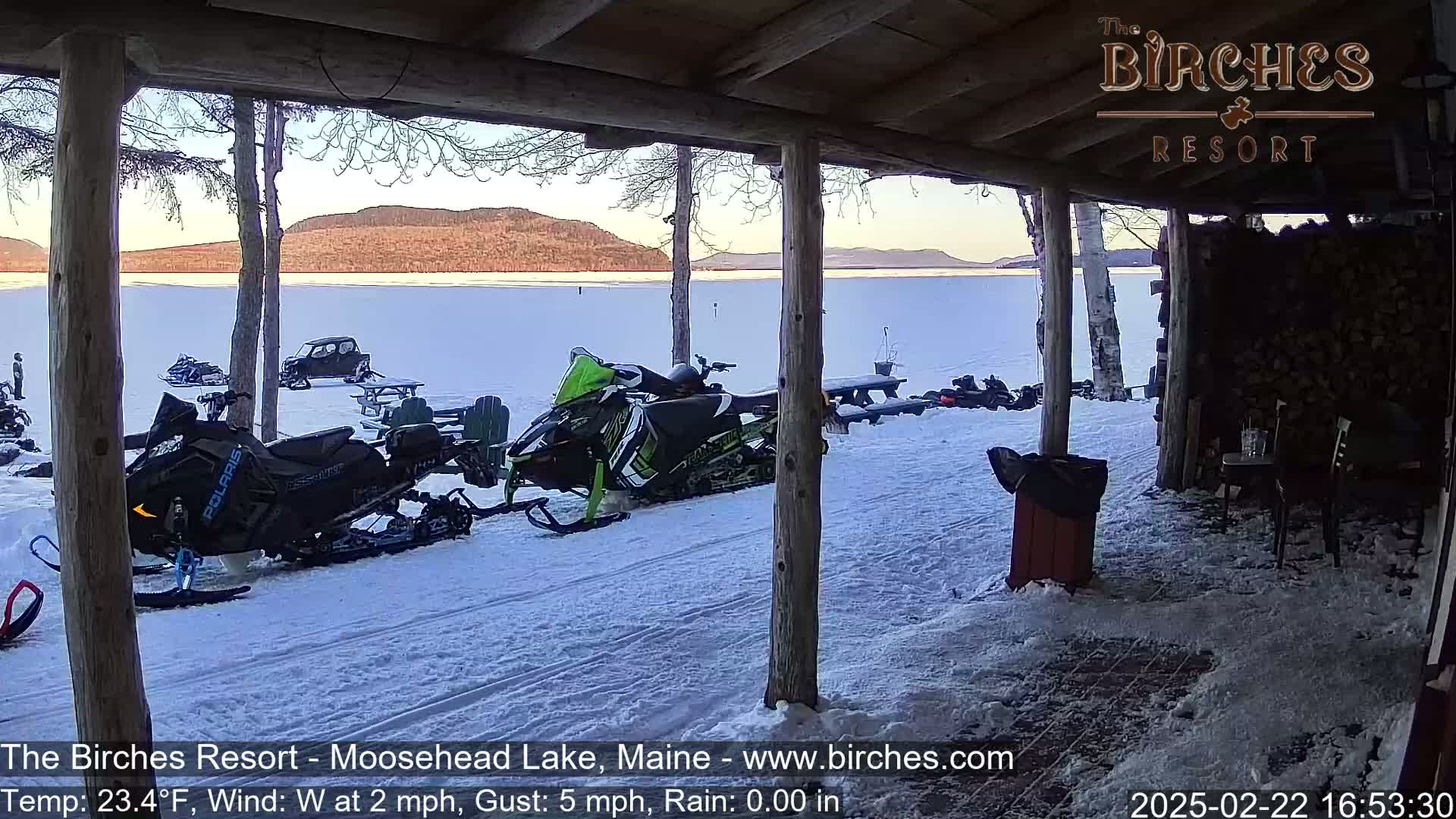From a snow-covered porch, several snowmobiles are parked near a frozen lake under a partly cloudy sky.