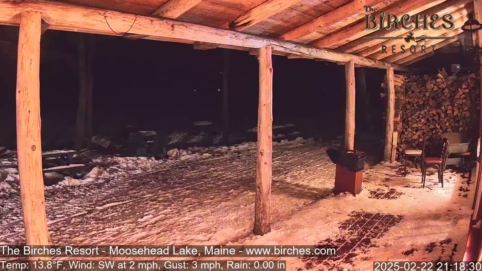 A snow-covered outdoor patio area is seen at night under a wooden-beam covered structure, with two chairs and a woodpile visible.