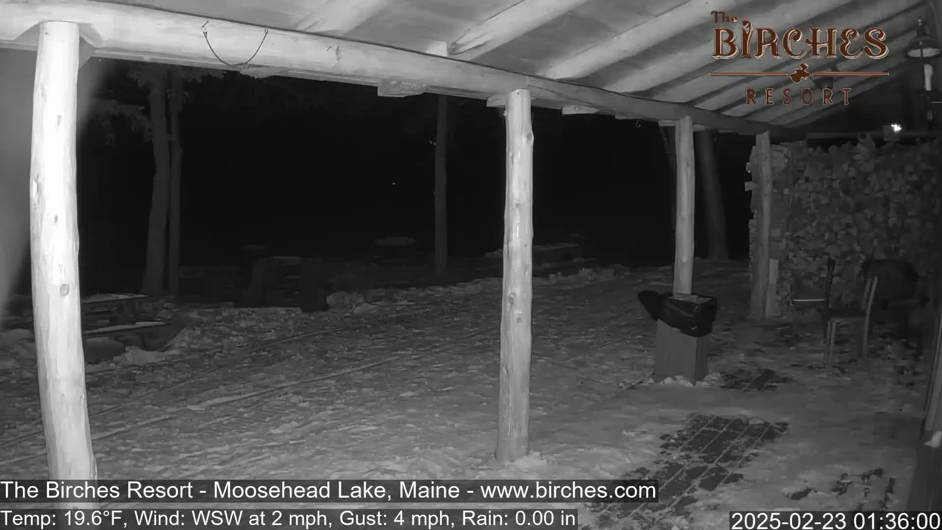 A snow-covered outdoor patio area under a wooden covered structure at night, with a slight wind and rain.