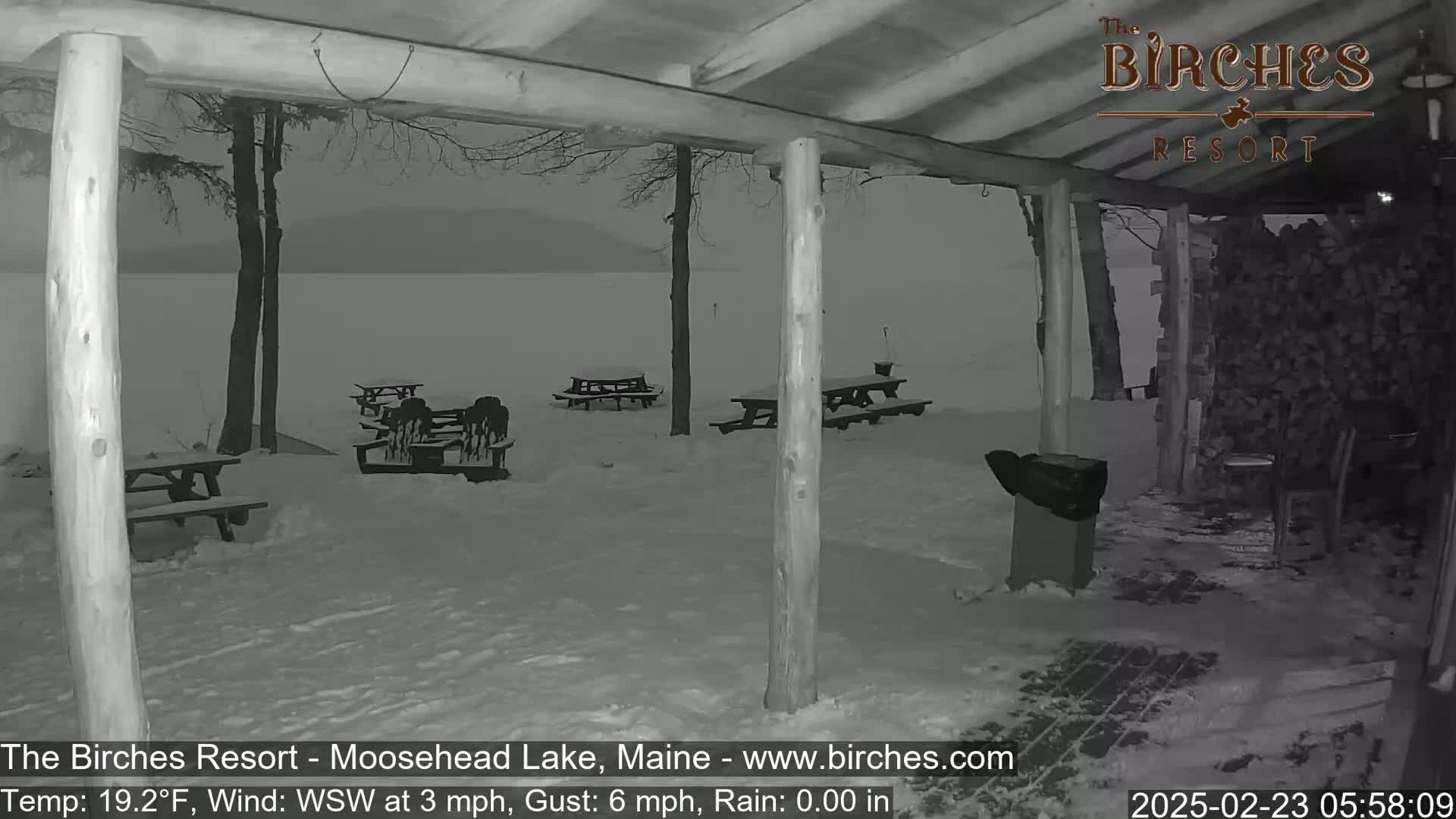A snow-covered outdoor patio area with picnic tables overlooks a lake under a gray sky, during a light rain.