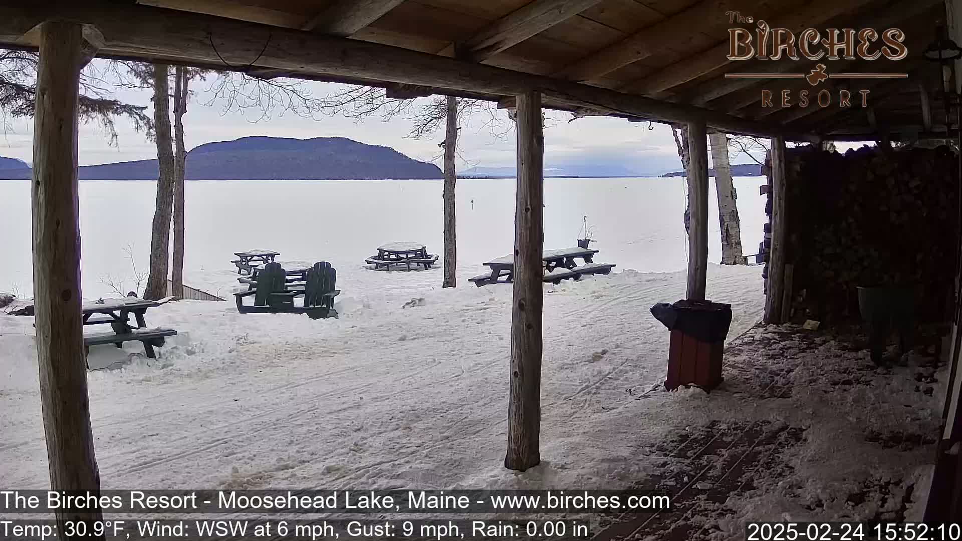 A snow-covered lakeside scene is viewed from a wooden porch, showing picnic tables and Adirondack chairs in the snow with a calm lake and distant mountains in the background.