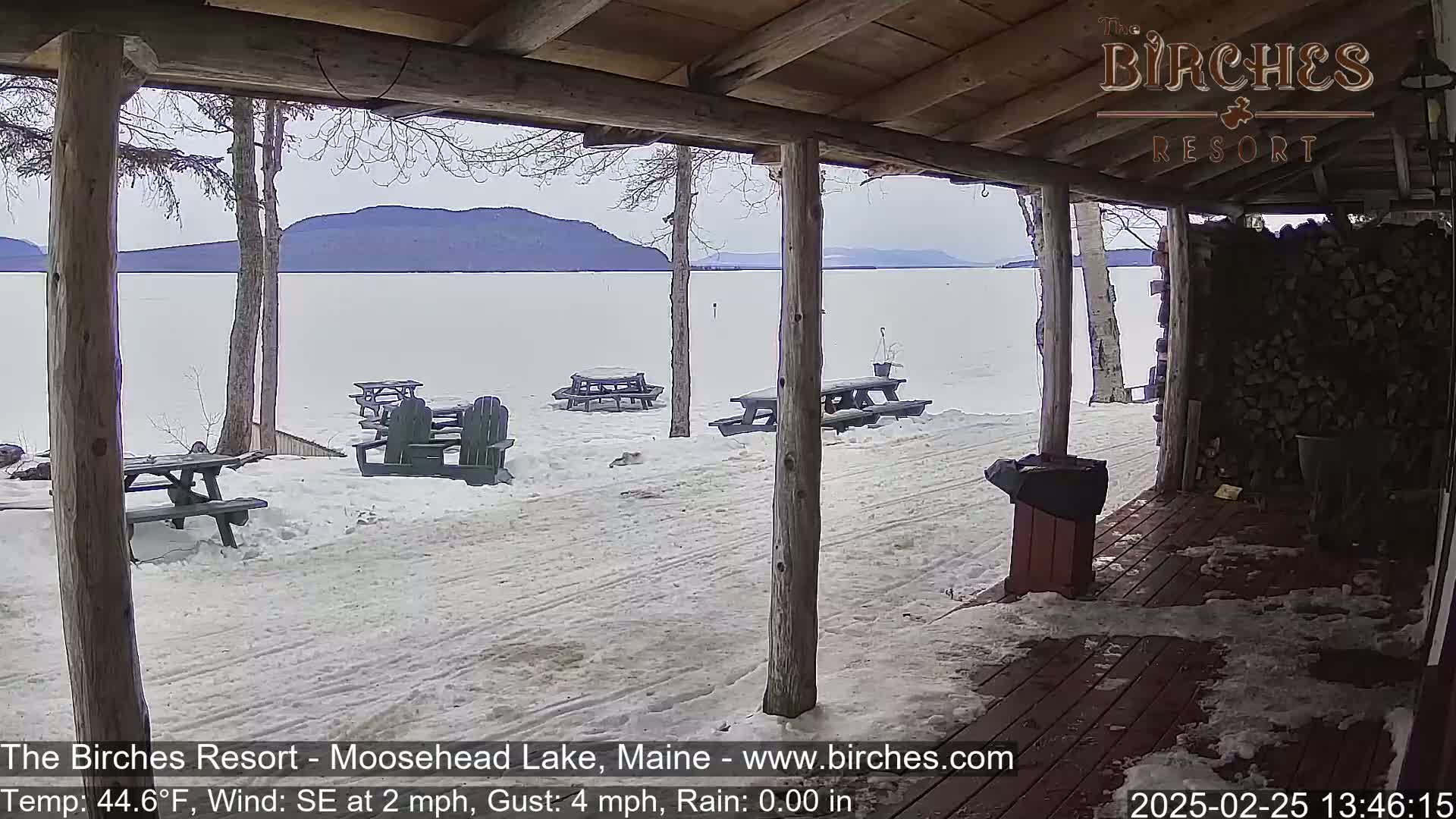 A snow-covered lakeside patio with picnic tables and chairs is viewed from a covered porch, with mountains visible in the distance under a cloudy sky.