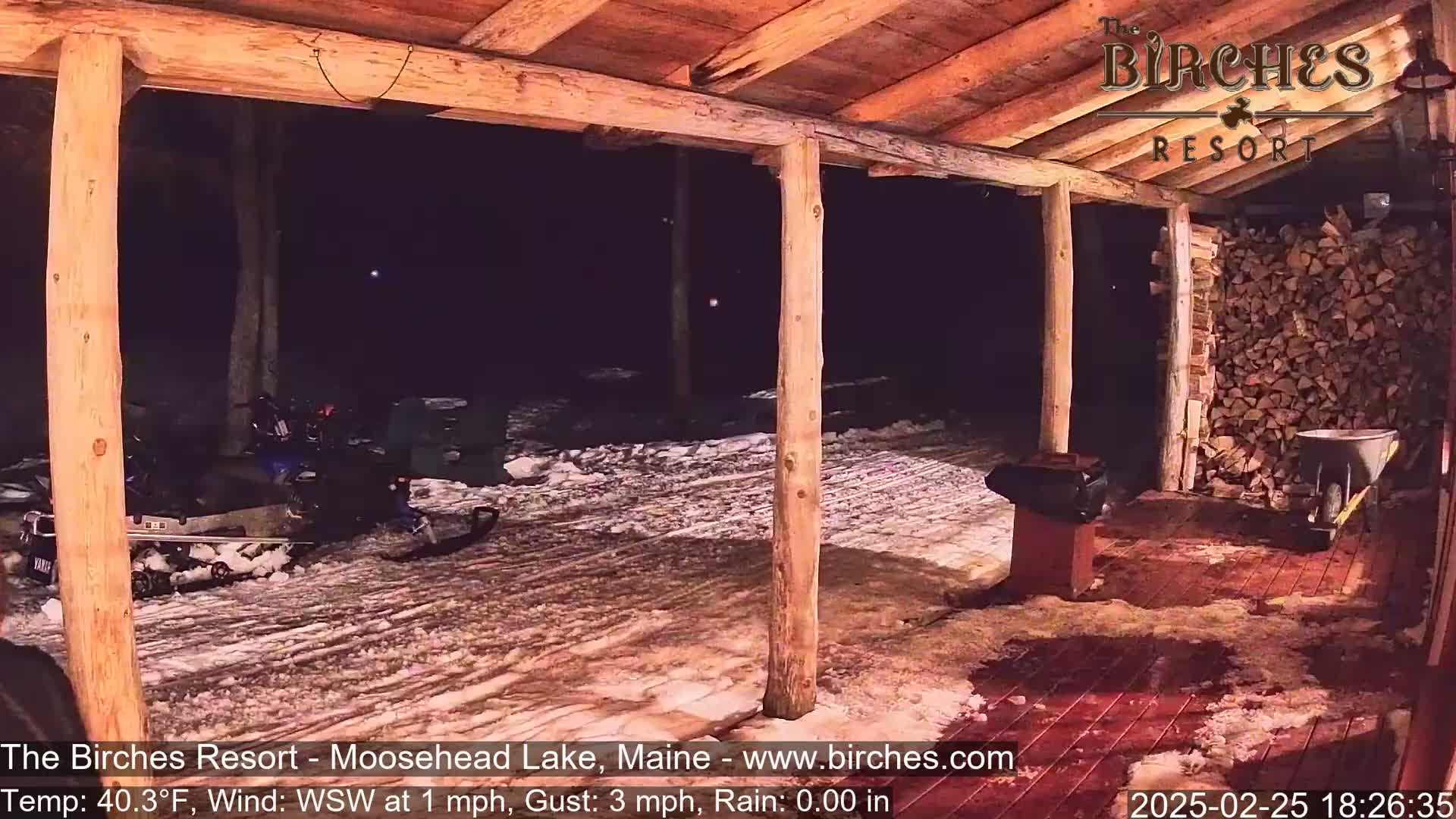 A snowmobile is parked outside a wooden porch at night in snowy conditions with a slight rain.