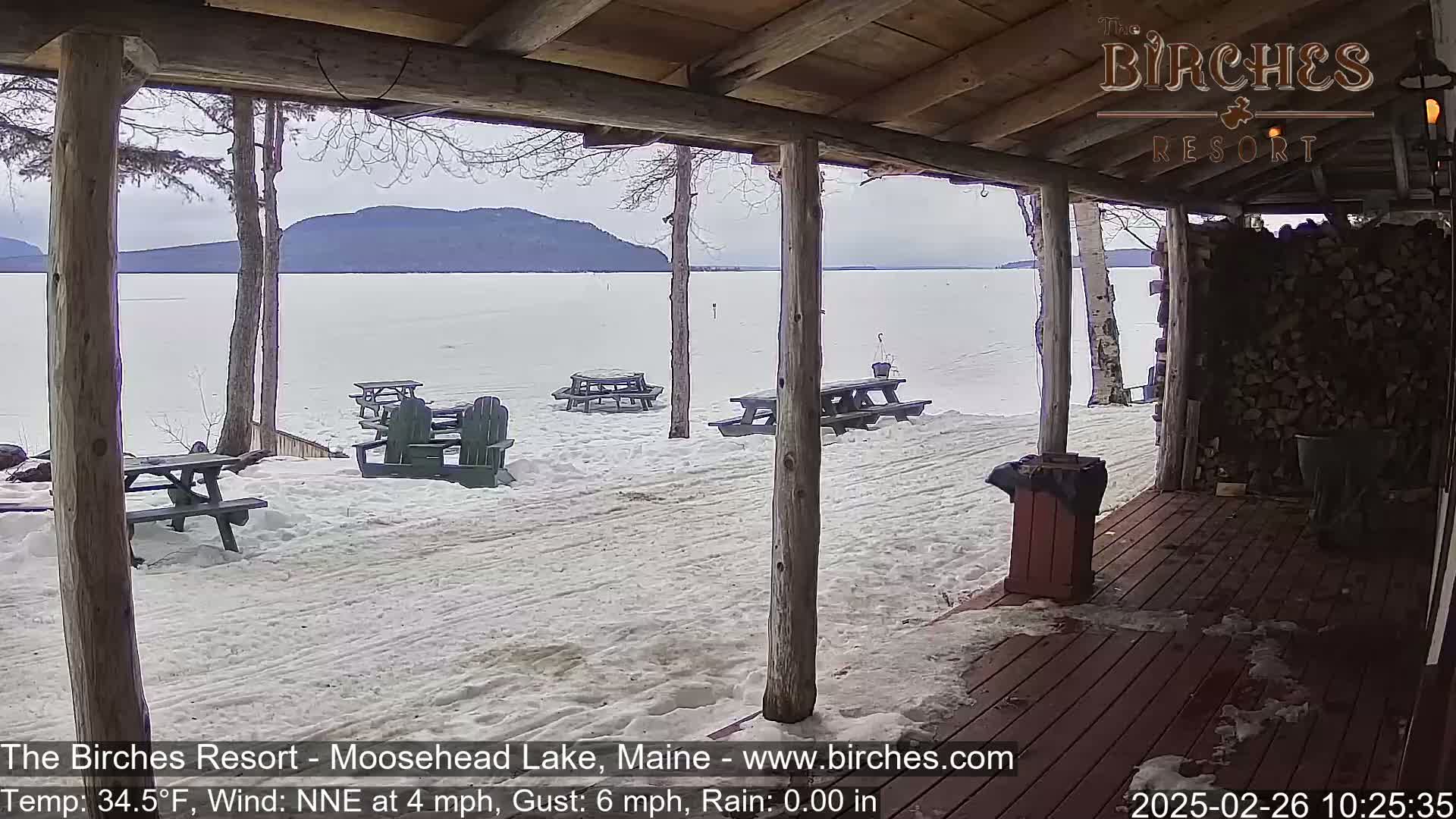 A snow-covered lakeside view is seen from a wooden porch with picnic tables and chairs, under a cloudy sky.