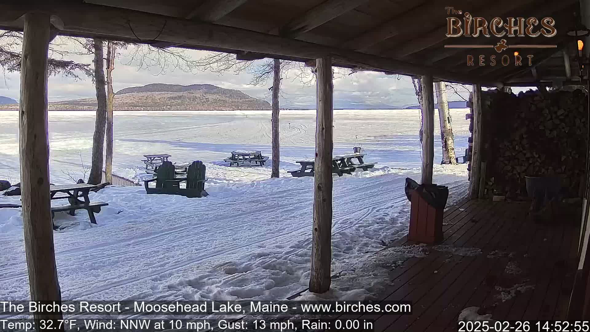 A snow-covered porch overlooks a partially frozen lake with distant mountains under a partly cloudy sky.
