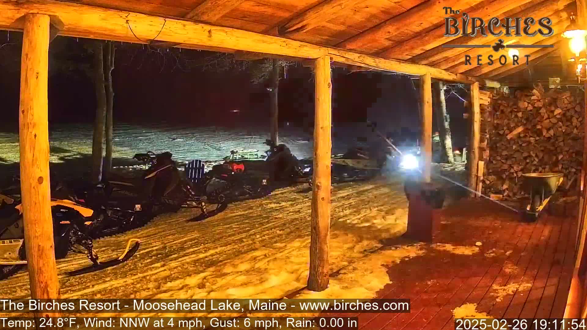 Several snowmobiles are parked outside a wood-framed porch at night in snowy conditions with a temperature of 24.8°F, a north-northwest wind at 4 mph gusting to 6 mph, and no rain.