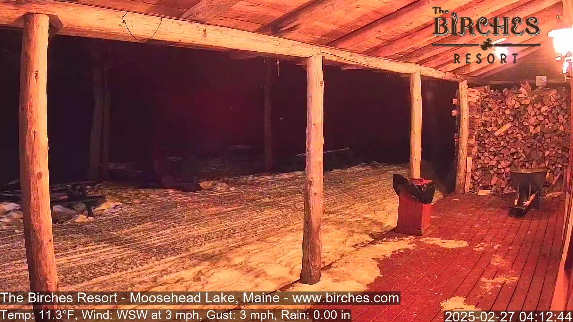 A snow-covered porch with wooden posts and a woodpile at night, illuminated by artificial light.