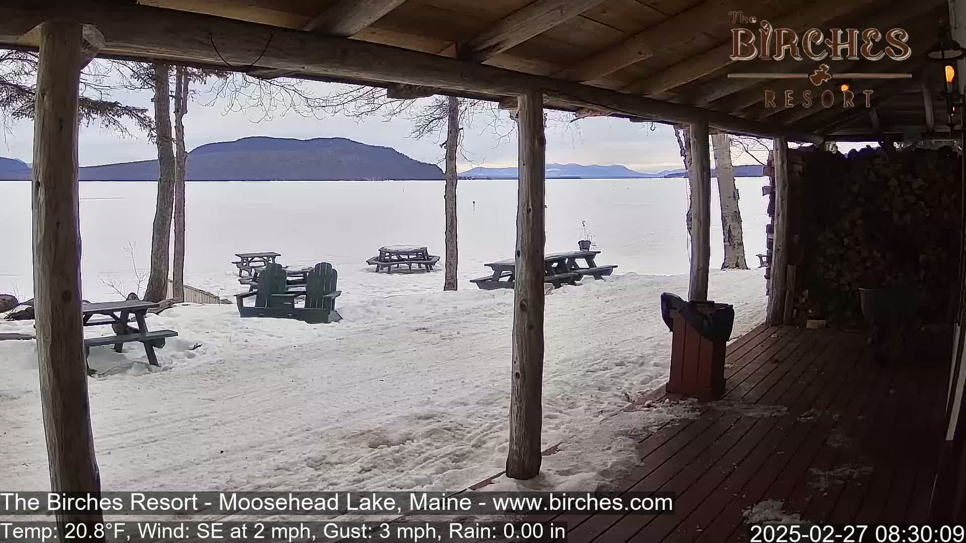 A snow-covered lakeside area with picnic tables and Adirondack chairs is viewed from a wooden porch on an overcast day.