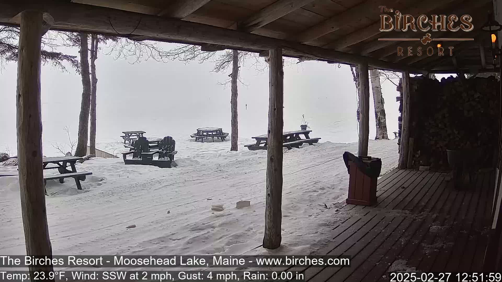 A snow-covered outdoor patio with picnic tables and chairs is seen from a covered porch during a light rain.