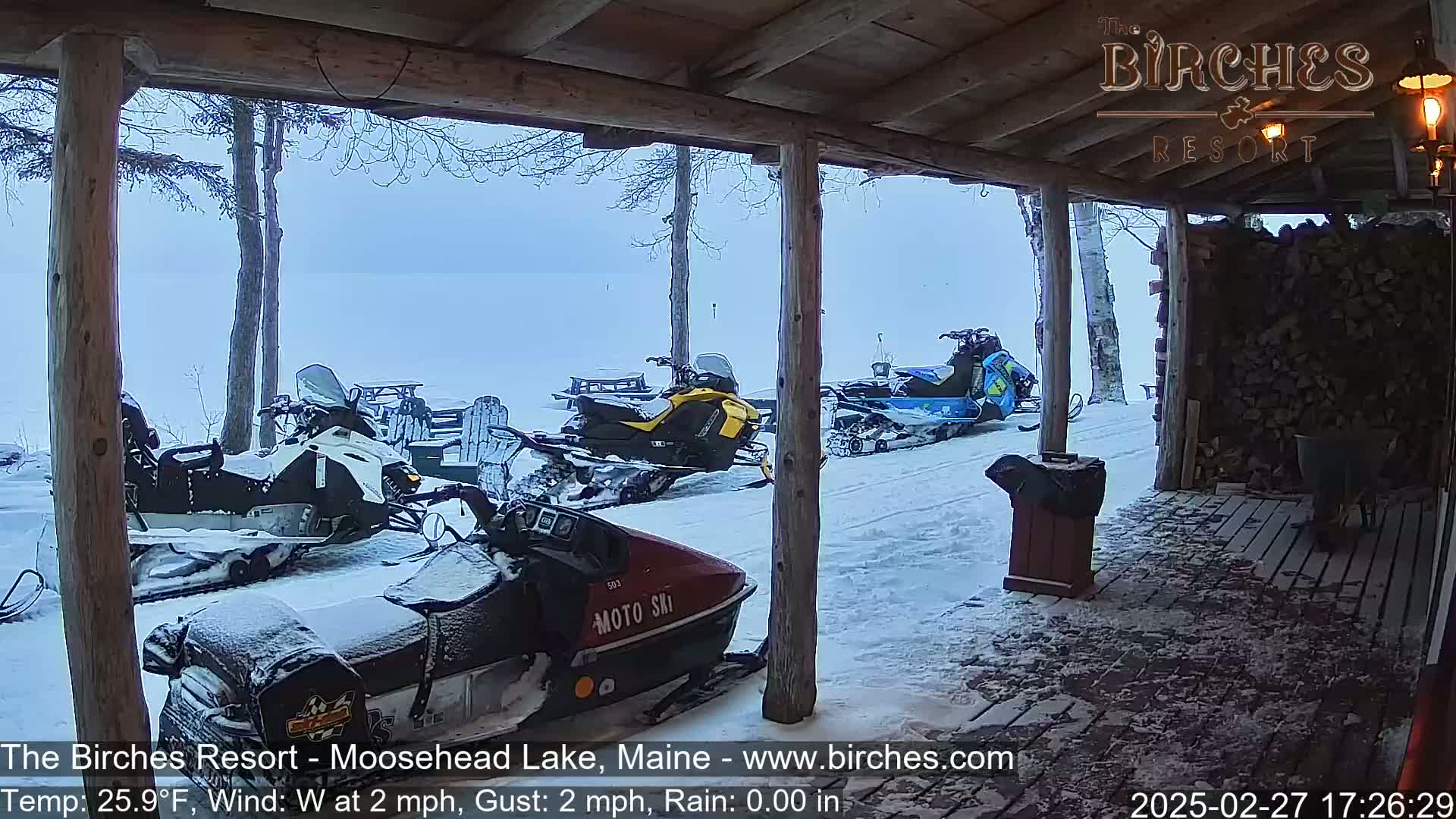 Several snowmobiles are parked in the snow outside a wooden porch on a hazy, snowy day with a light westerly wind.