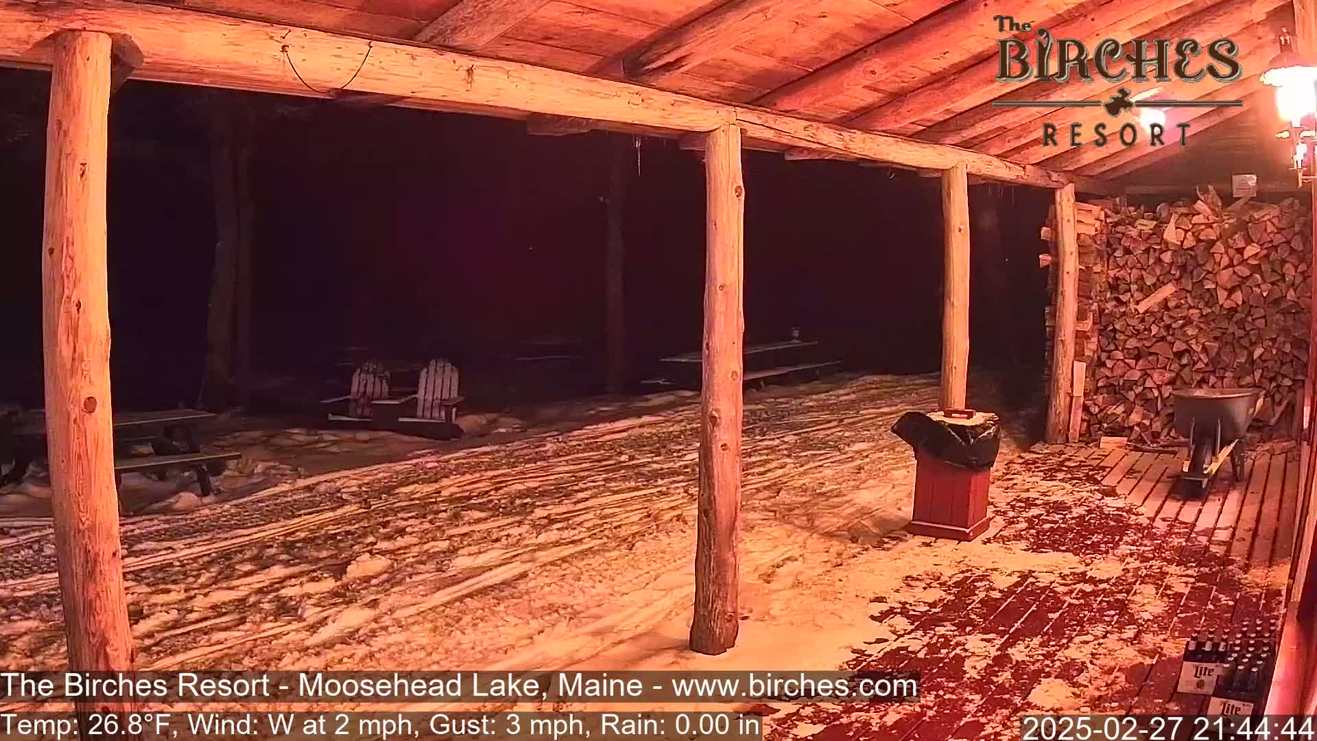 A snow-covered outdoor patio area with wooden posts and picnic tables under a wooden shelter at night, with a light wind and no rain.