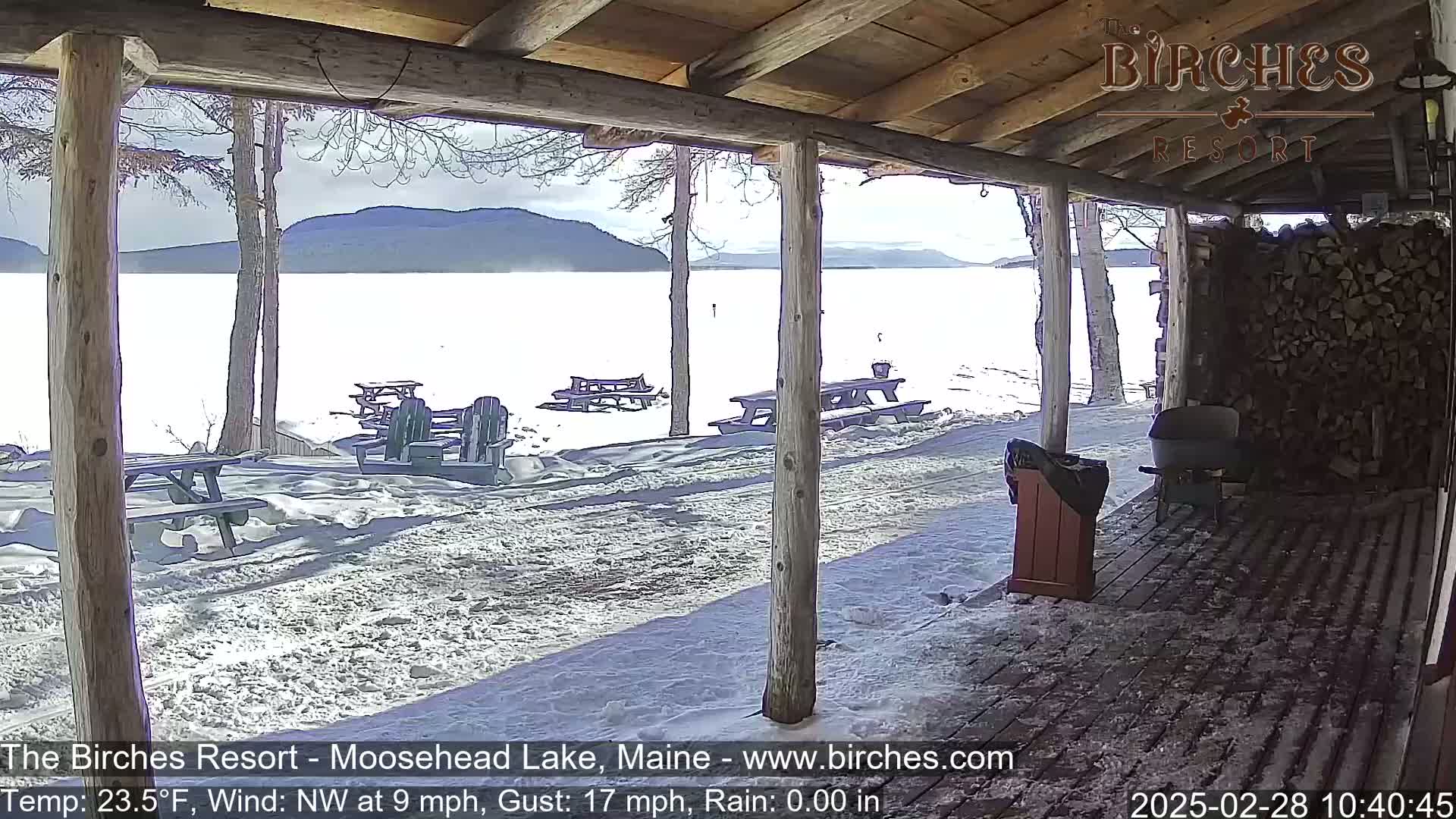 A snow-covered porch overlooks a frozen lake and distant mountains under a partly sunny sky.