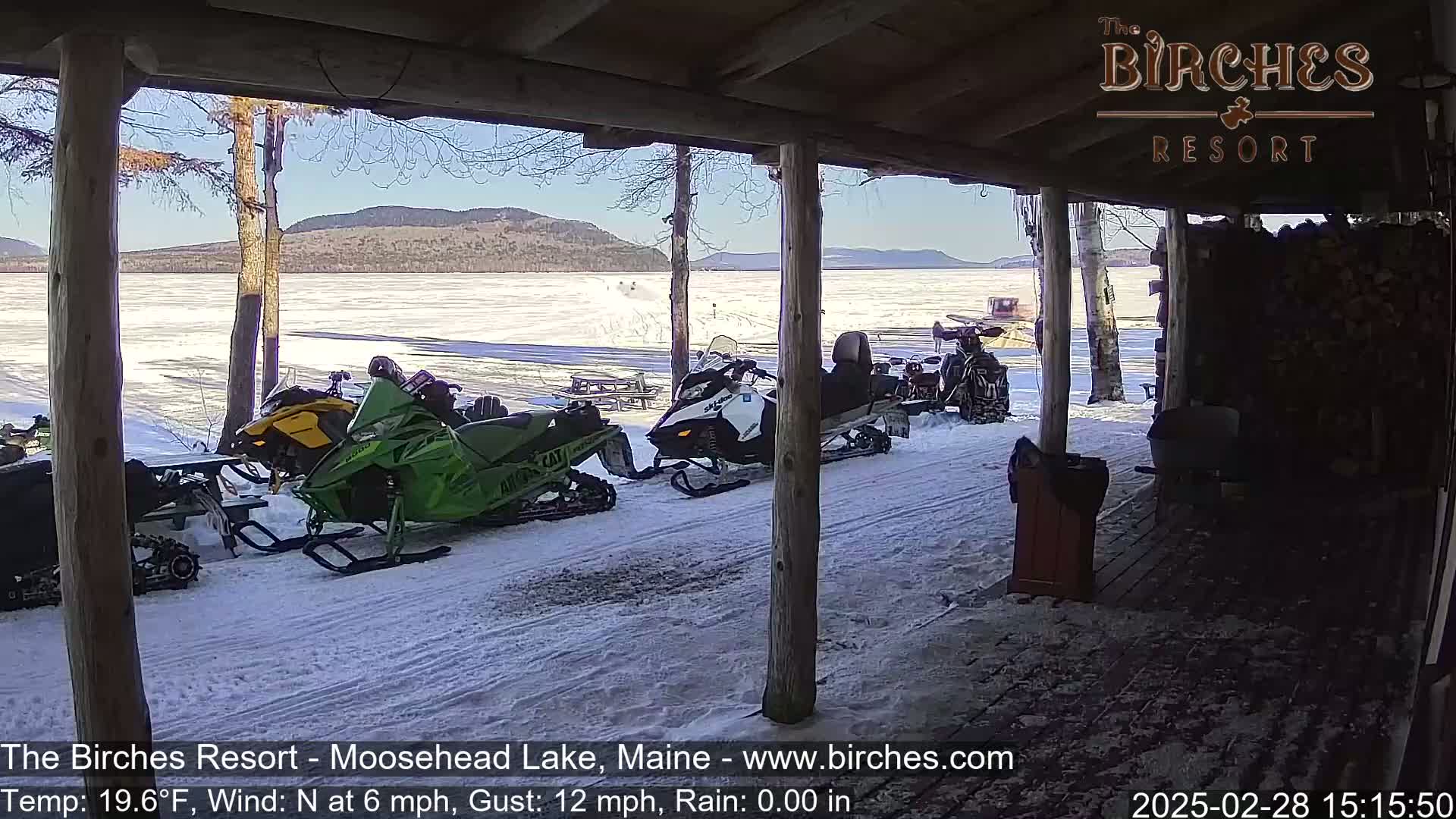 Several snowmobiles are parked outside a wooden structure overlooking a frozen lake under a sunny sky.