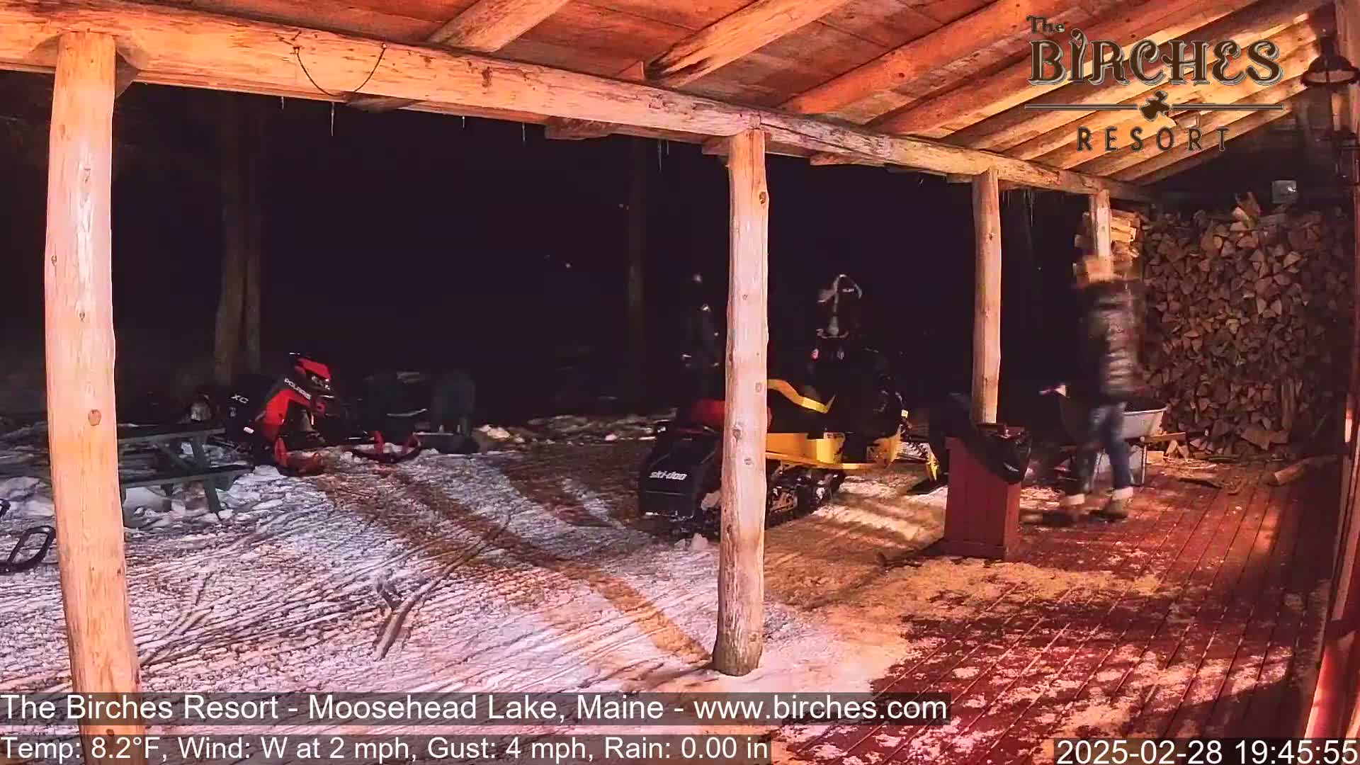 A snow-covered outdoor area with several snowmobiles parked under a wooden structure at night, with a person walking nearby.