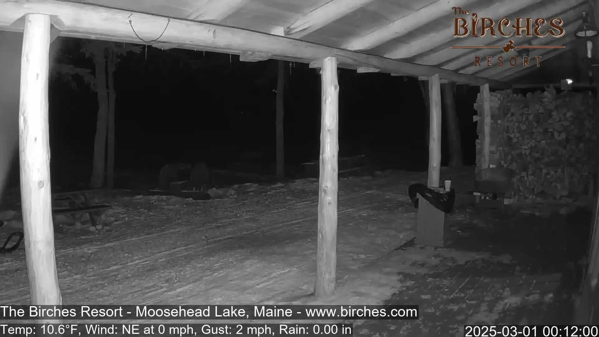 A nighttime, snowy scene is visible from under a covered porch, showing a woodpile and a dark yard beyond.
