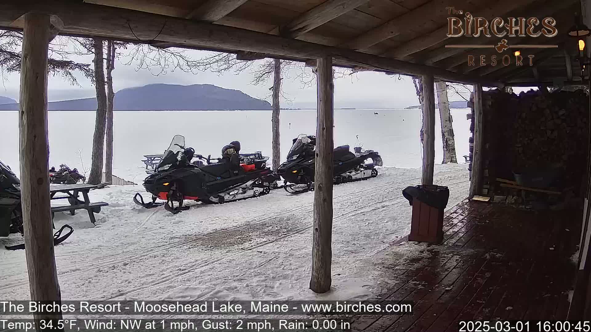Several snowmobiles are parked outside a wooden structure overlooking a snow-covered lake and distant mountains under a cloudy sky.