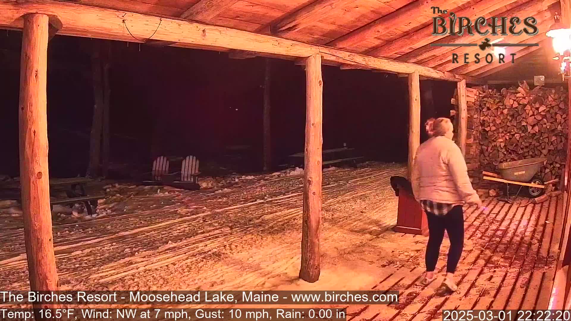 A person walks on a snow-covered porch at night under a wooden structure, with a pile of firewood and Adirondack chairs visible in the background.