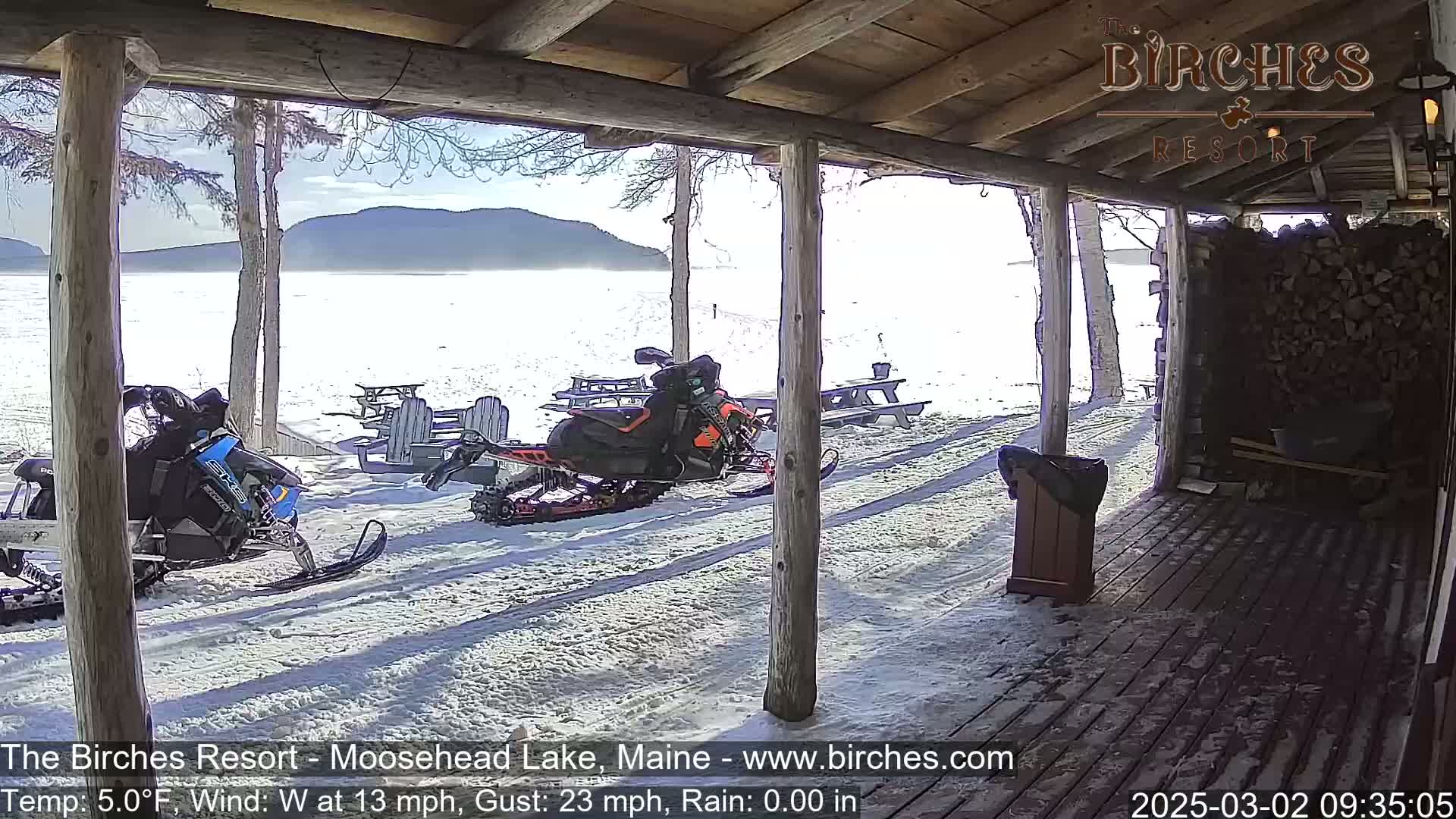 Two snowmobiles are parked on a snow-covered porch overlooking a calm, snow-covered lake and distant mountains under a sunny sky.