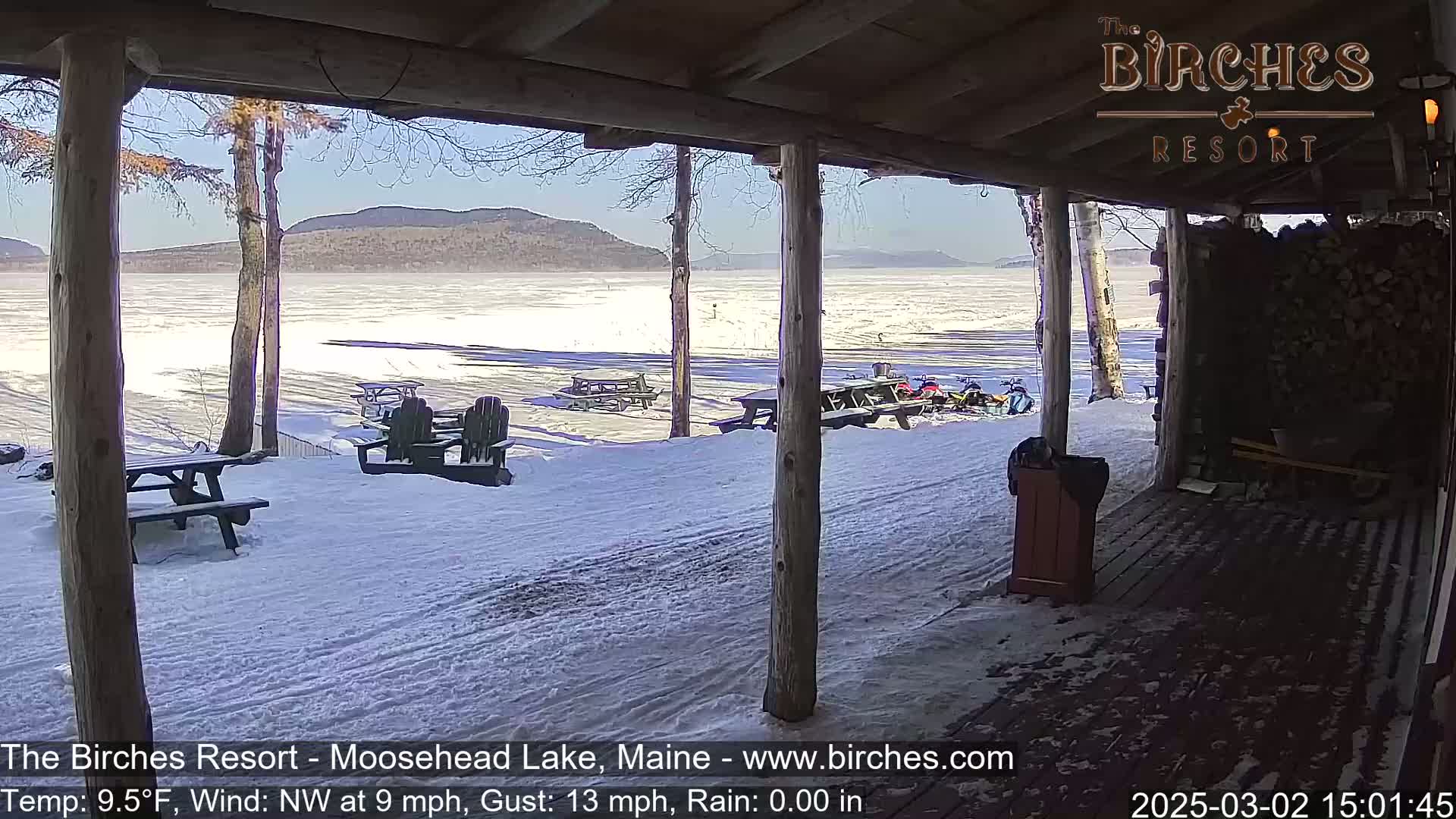 A snow-covered lakeside view is seen from a covered porch, with picnic tables and snowmobiles visible in the distance under a sunny sky.