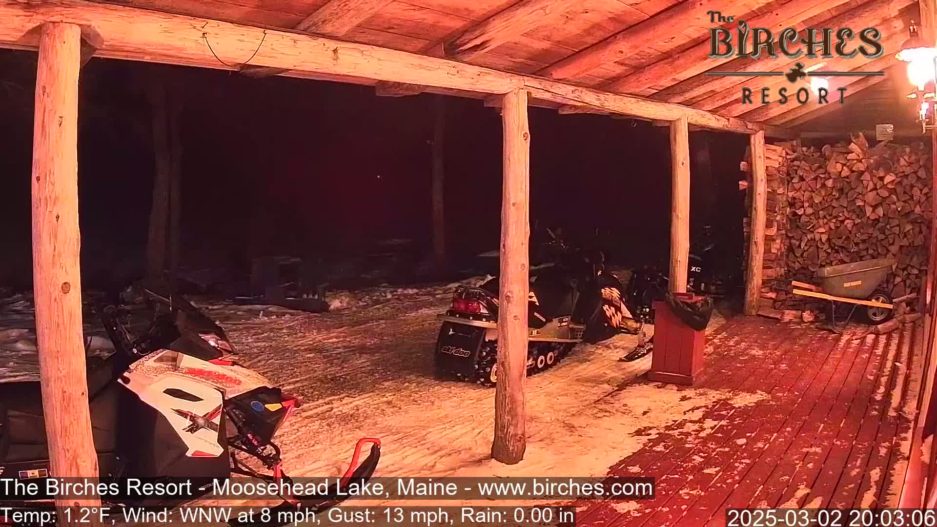 Under a covered porch at night in snowy conditions, two snowmobiles are parked near a woodpile and a wheelbarrow.