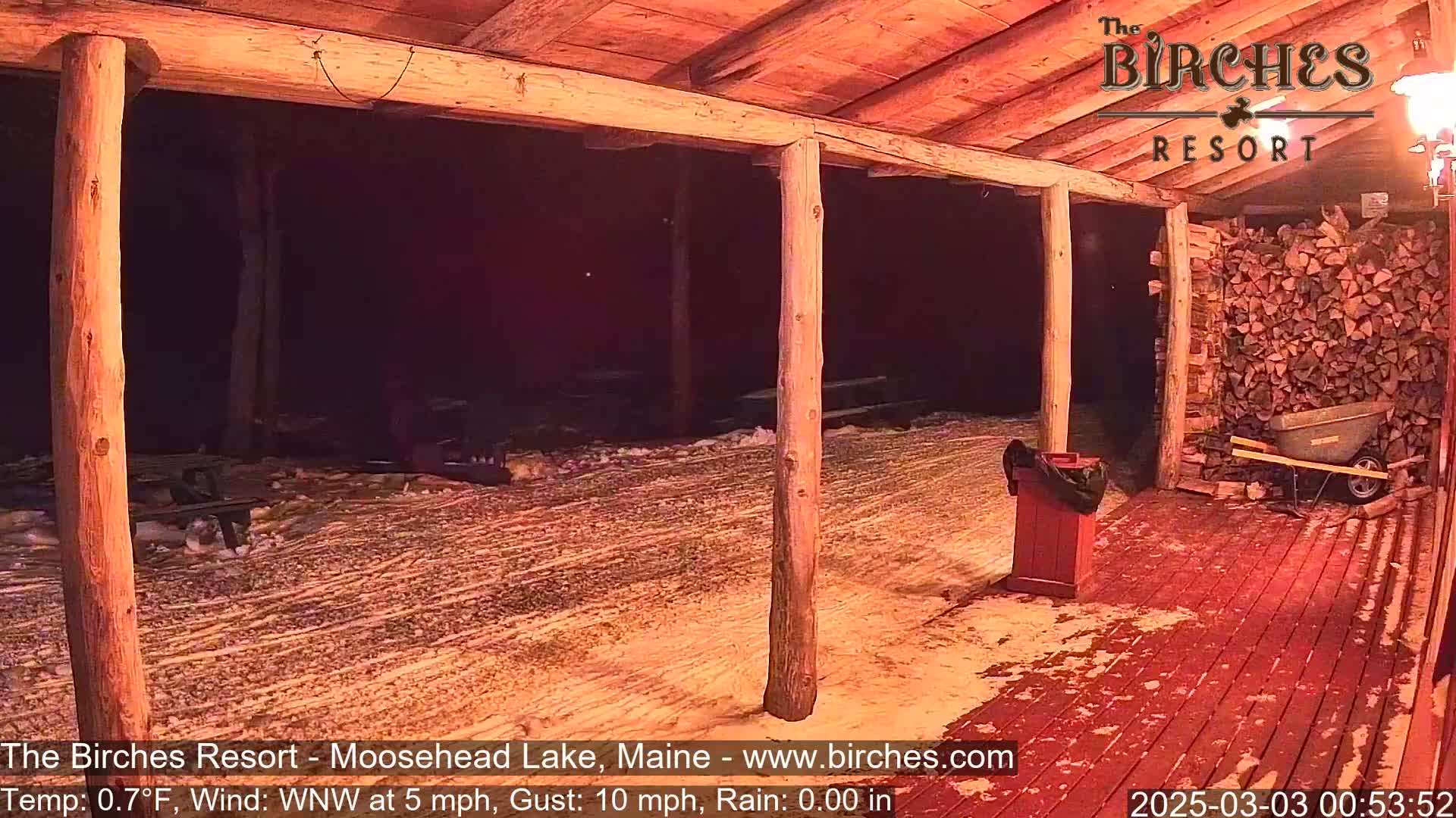 A snow-covered outdoor patio with wooden posts and a woodpile is visible at night under a partially covered structure; a wheelbarrow sits near the woodpile.