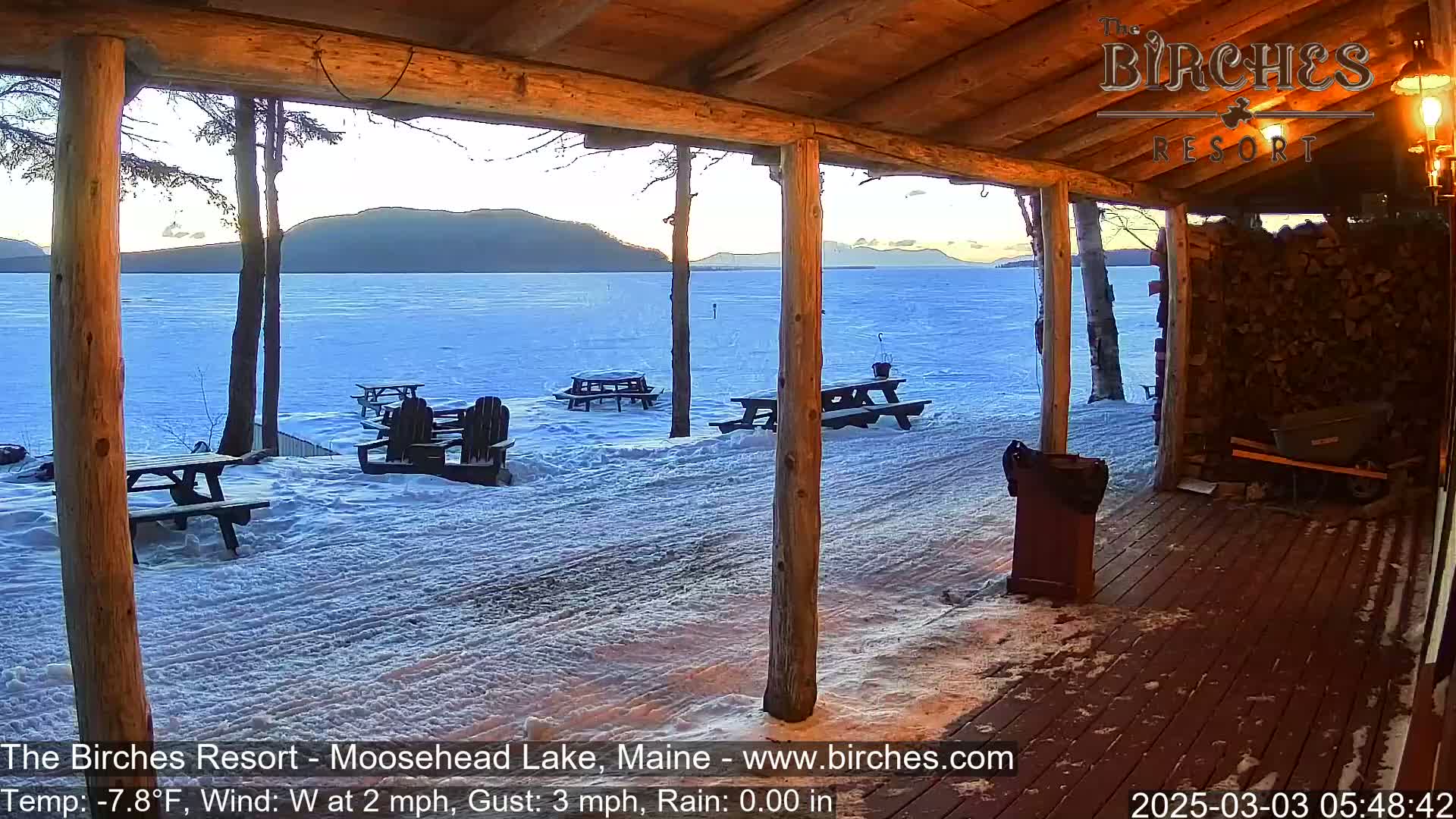 From a wooden porch overlooking a snow-covered lake and distant mountains, several empty picnic tables sit in the snow under a twilight sky.