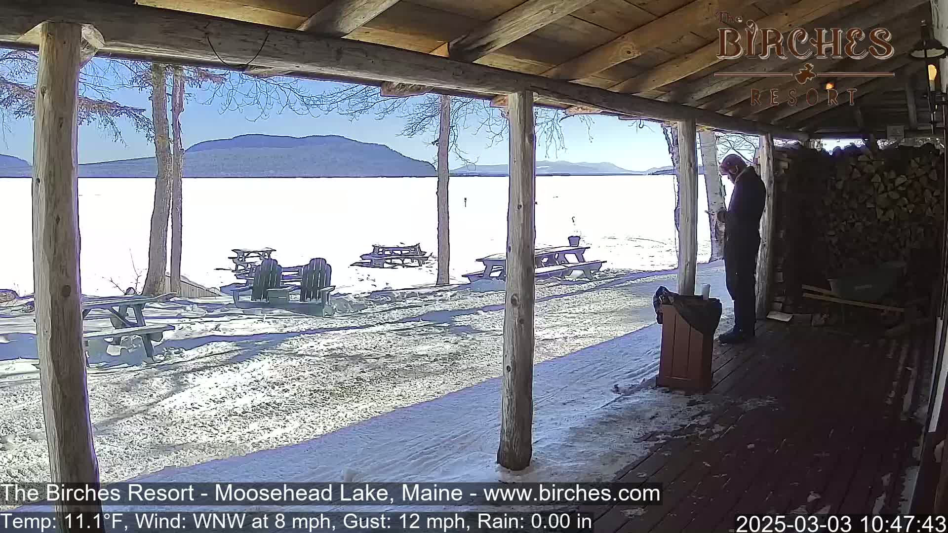 A person stands on a snow-covered porch overlooking a frozen lake and distant mountains under a clear, sunny sky.