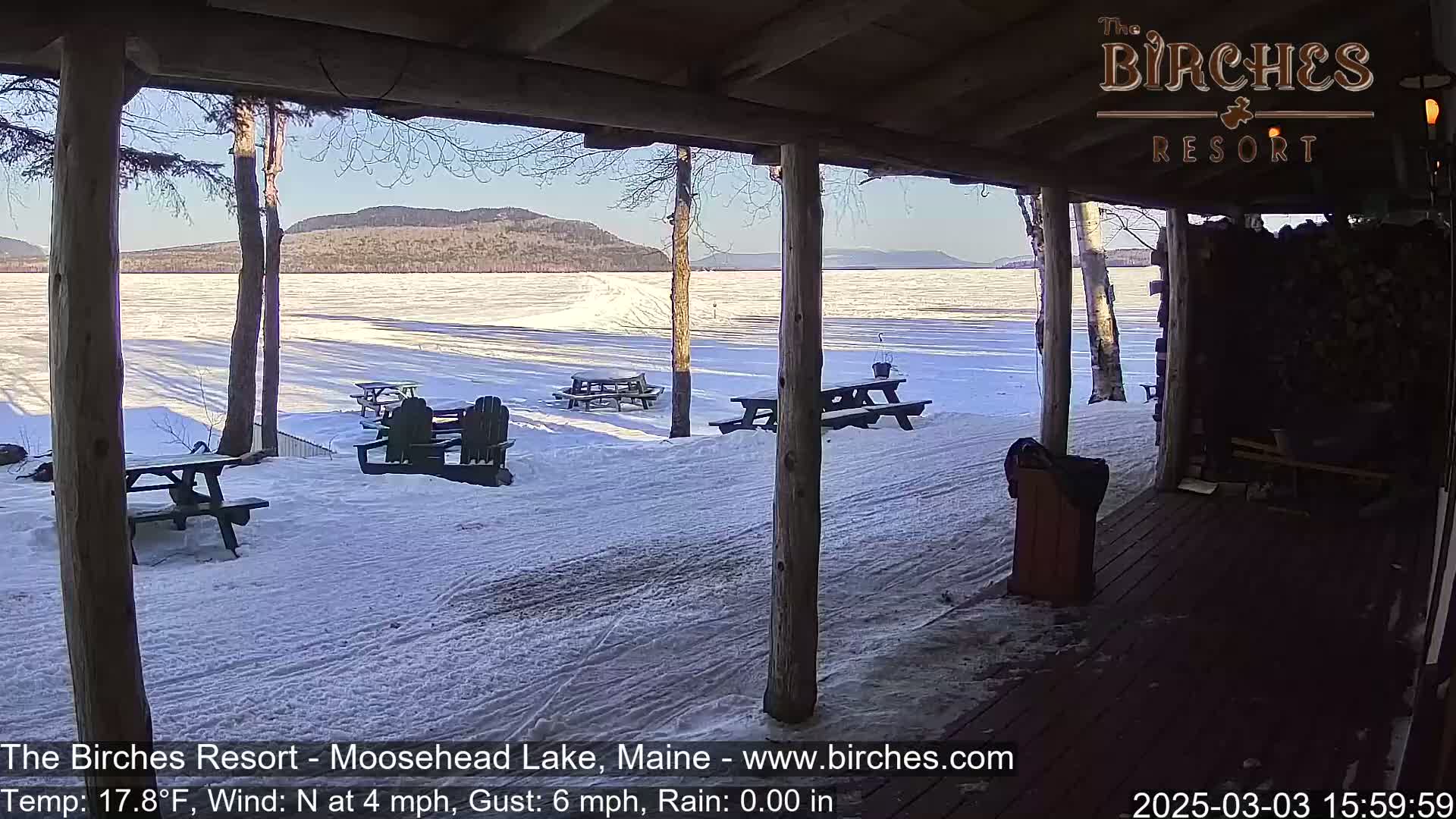 A snow-covered frozen lake is viewed from a wooden porch with picnic tables and chairs, under a sunny sky and a light north wind.